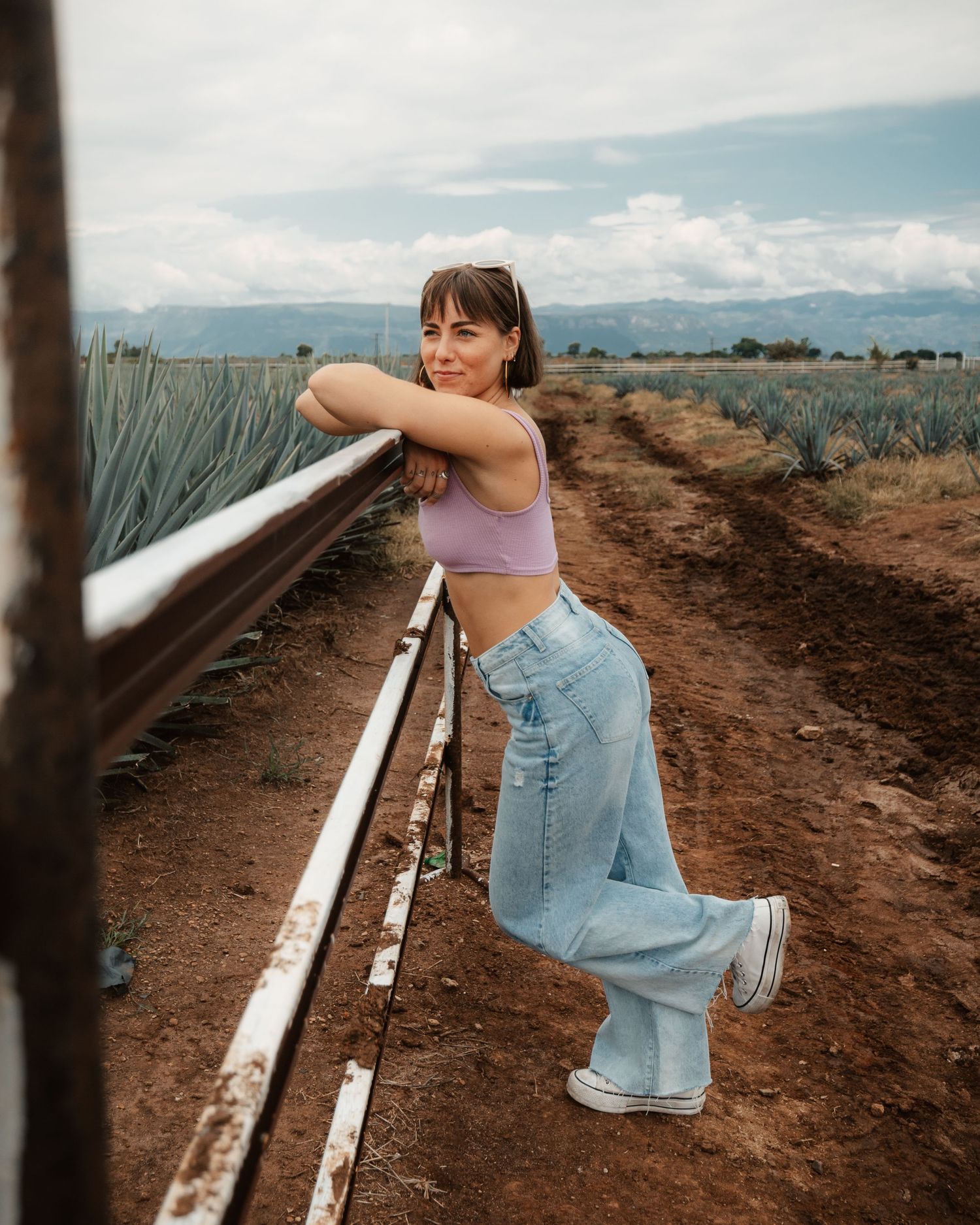 Woman in lavender crop top and wide-leg jeans leaning on a fence rail in an agave field.