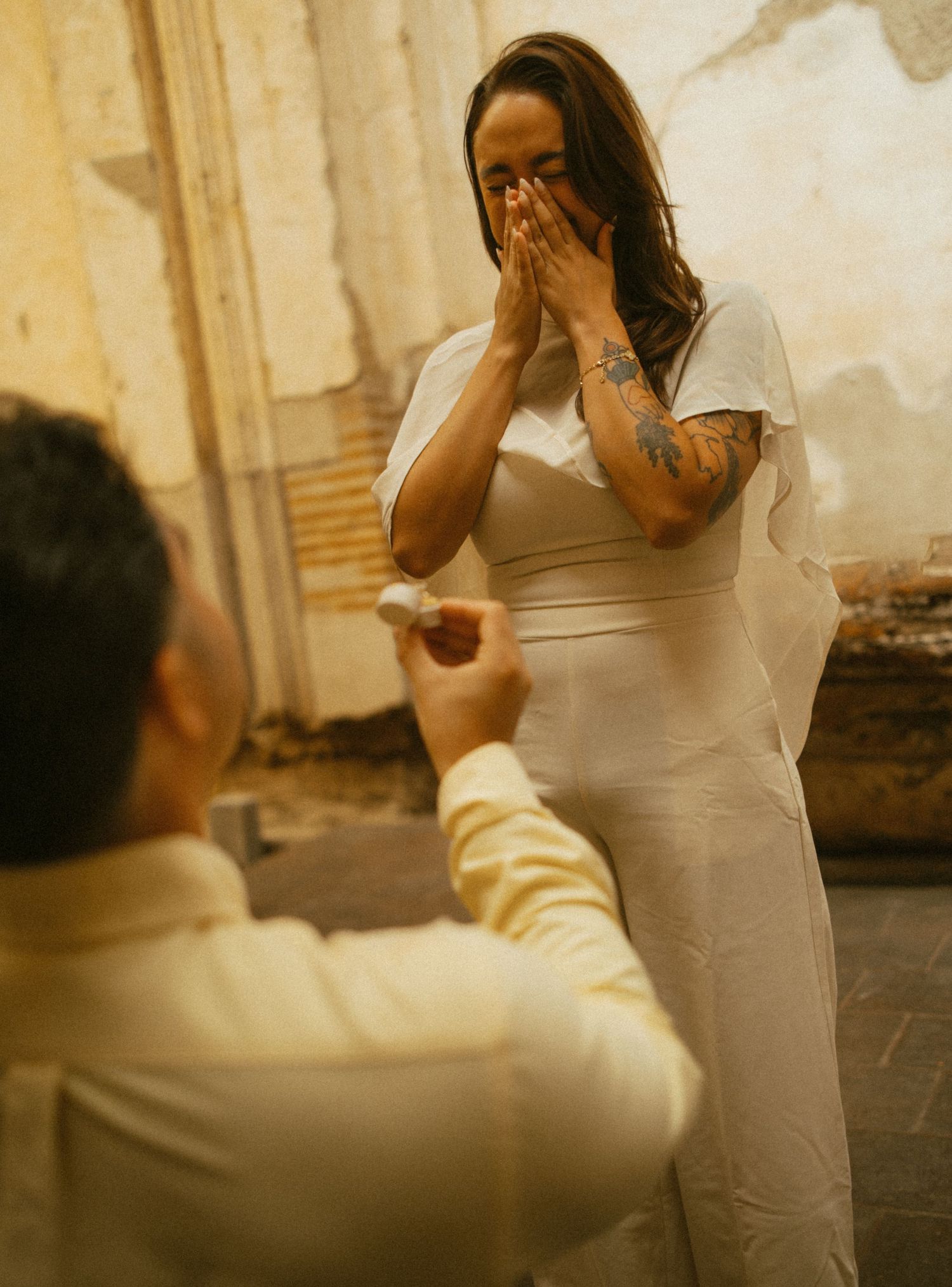 Man proposing to emotional woman in white dress with tattoos, hands covering her face in surprise, outdoor setting.