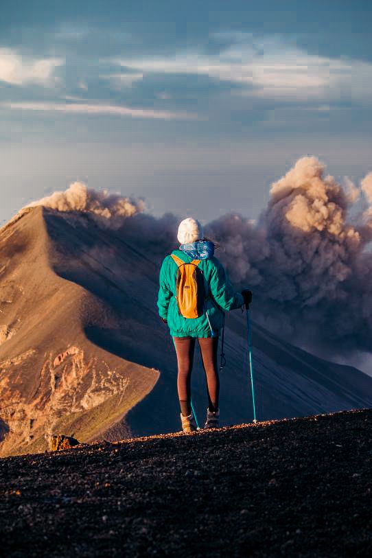 Hiker with yellow backpack and trekking poles stands on volcanic ridge watching eruption clouds rise.