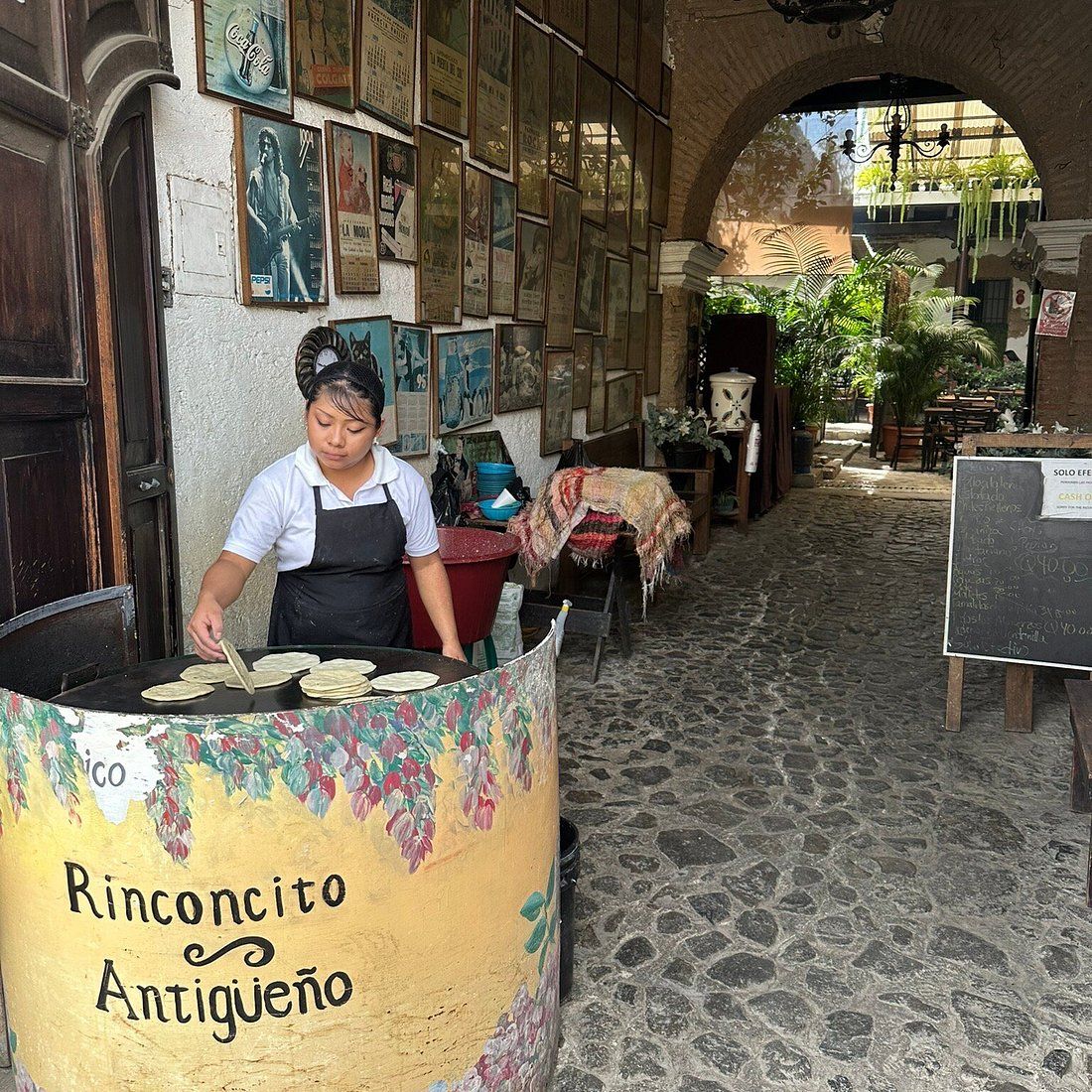 Woman cooking tortillas at Rinconcito Antigueño, a traditional Guatemalan street food stall in a cobblestone alley.