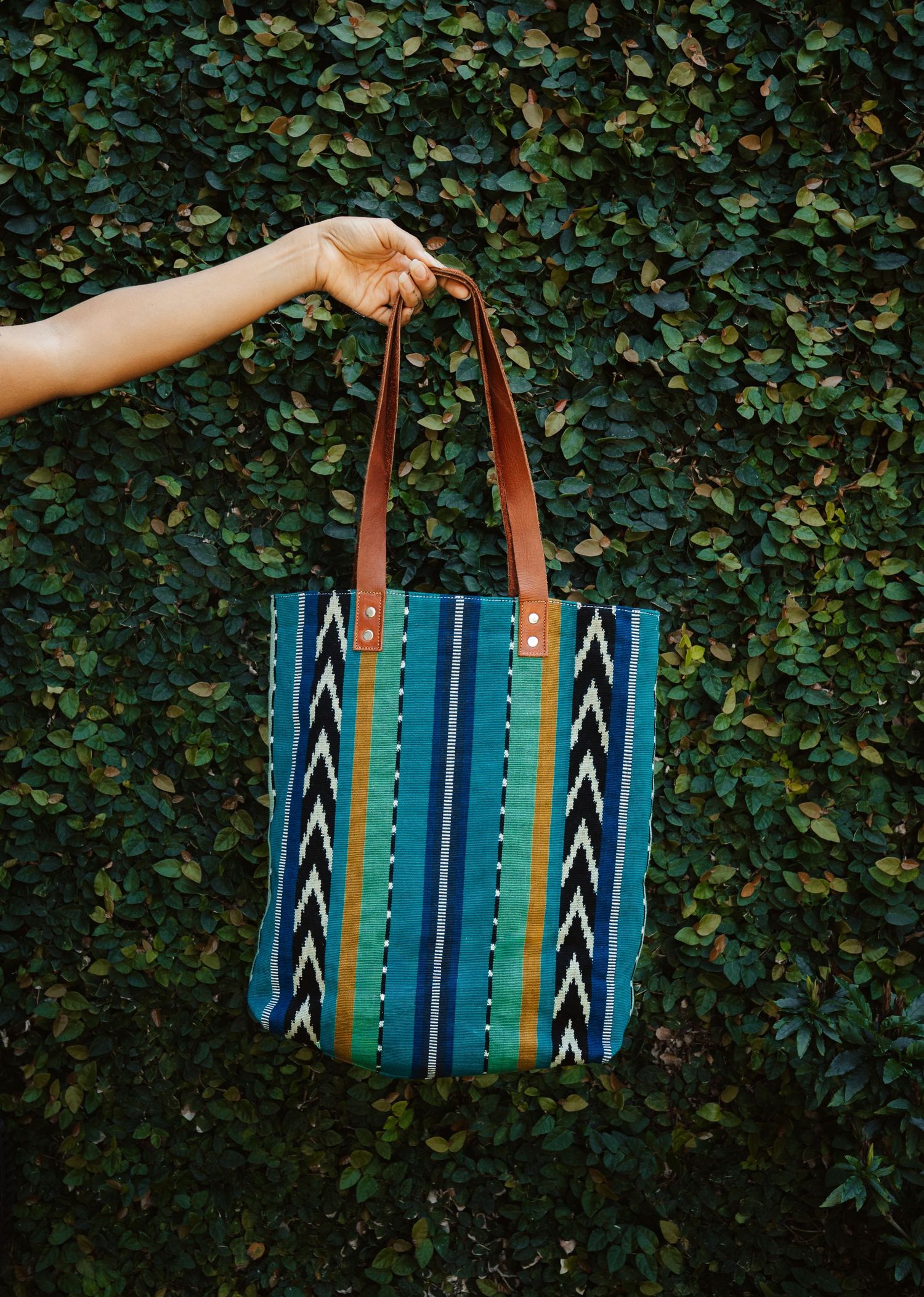 Colorful striped tote bag with brown leather handles held up against a green ivy wall.