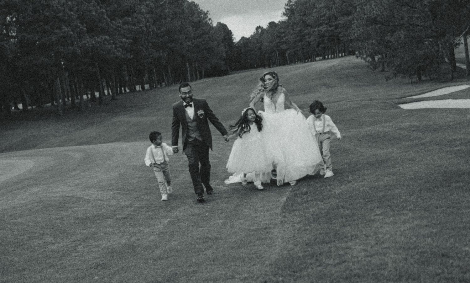 Black and white wedding photo of bride and groom walking with children on a lush golf course lined with tall trees.