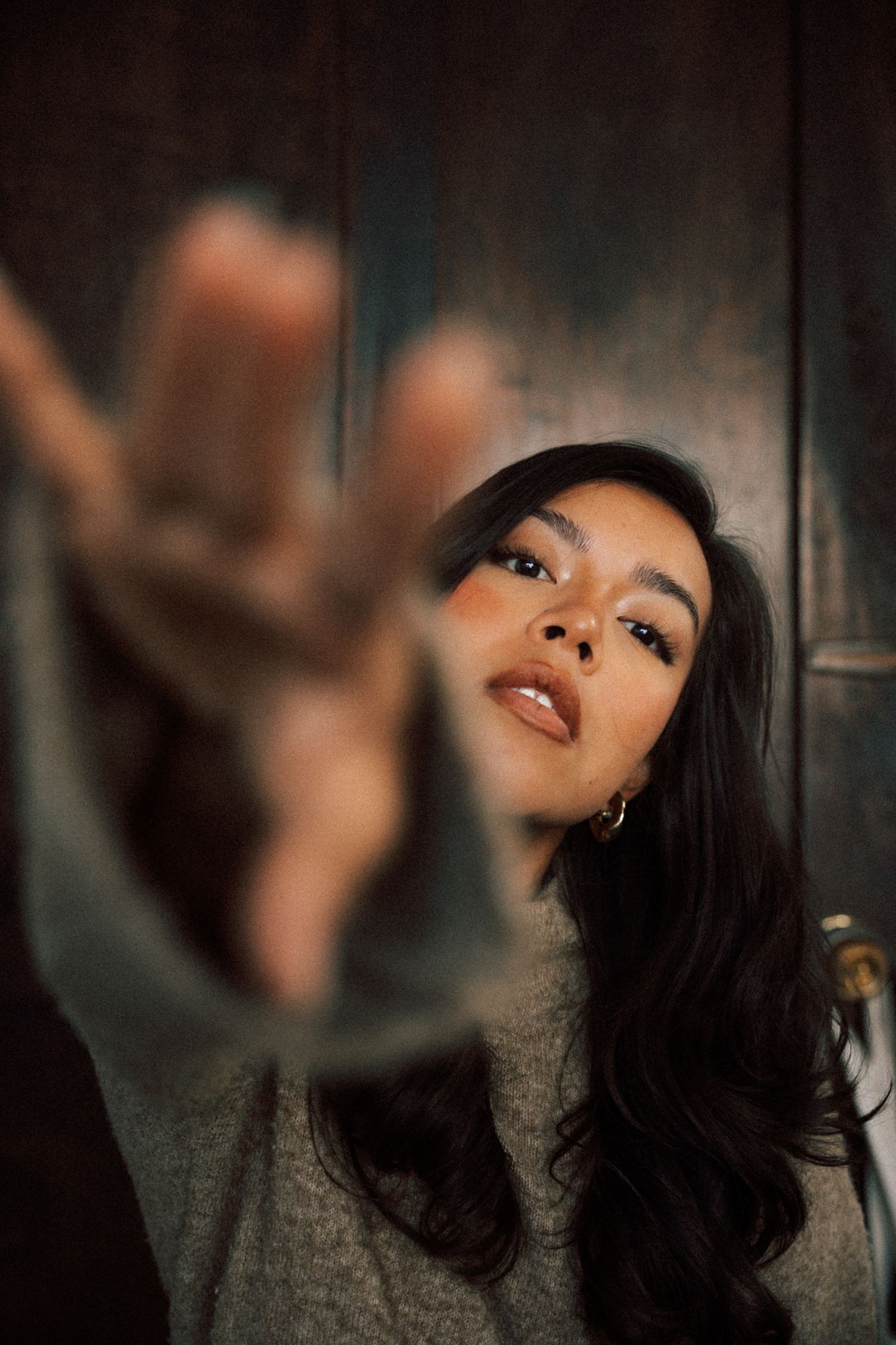 Young woman with long dark hair reaches toward camera with blurred hand in moody portrait shot.