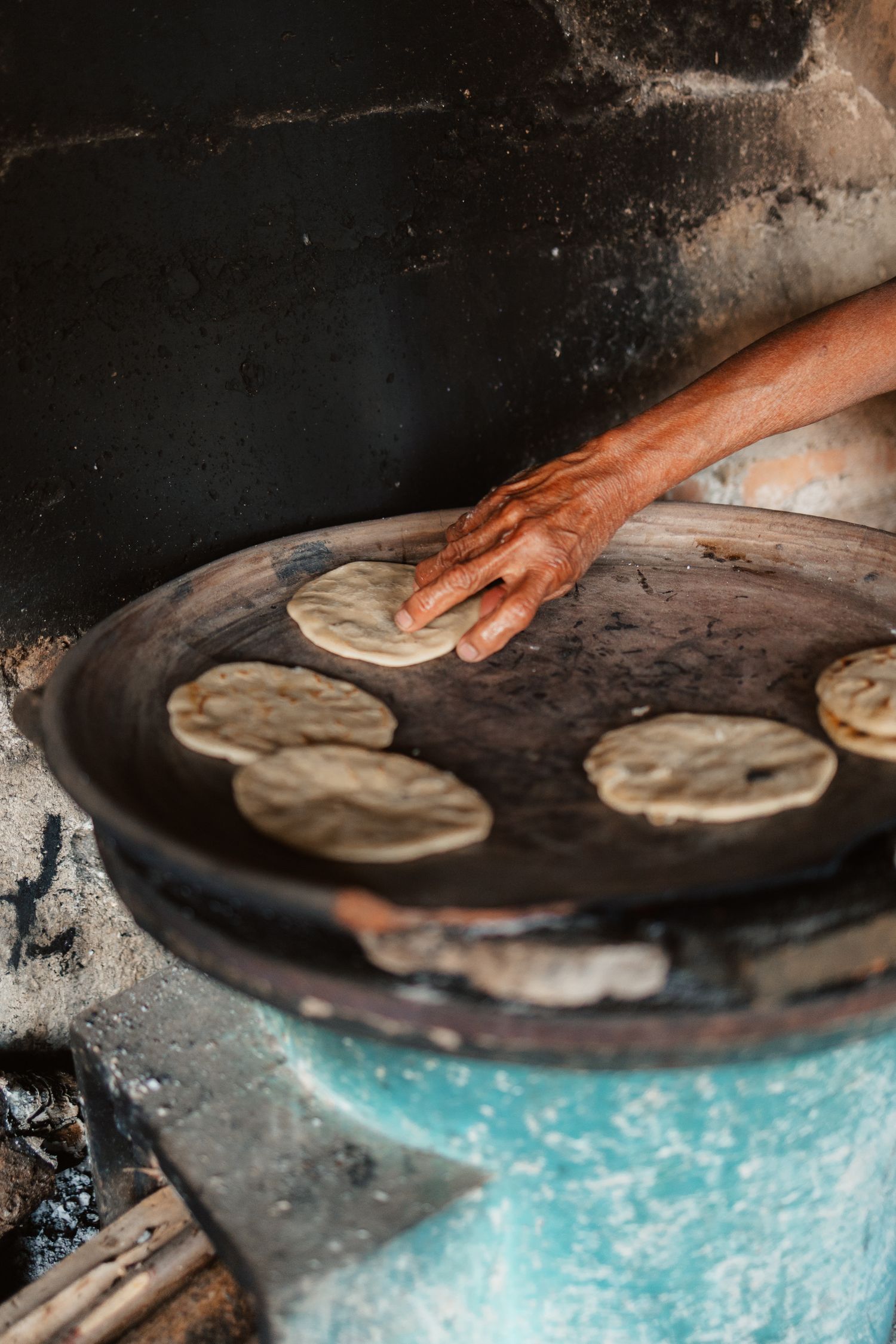 A hand placing raw tortillas on a traditional comal griddle over an open fire in a rustic kitchen.