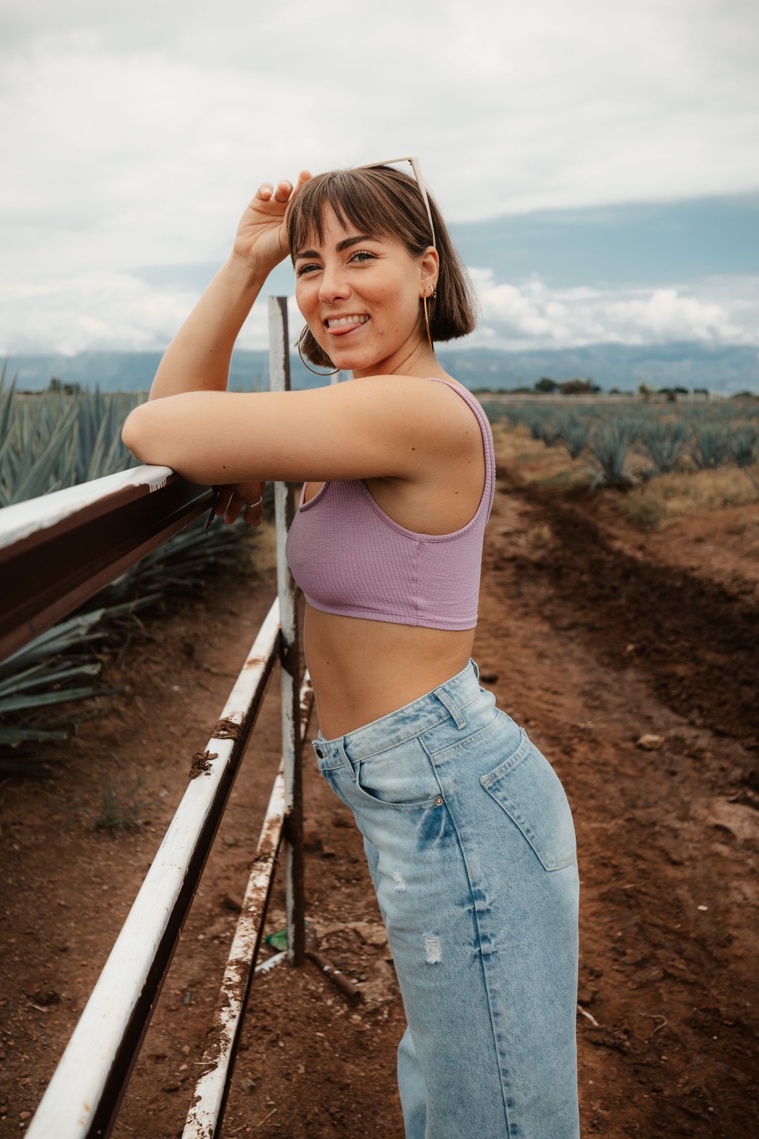 Smiling young woman in purple crop top and jeans leaning on fence in an agave field.