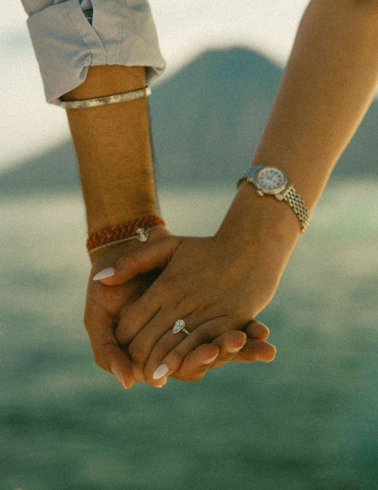 Couple holding hands outdoors, her hand wearing a diamond engagement ring and silver watch, with a blurred mountain and sea behind.