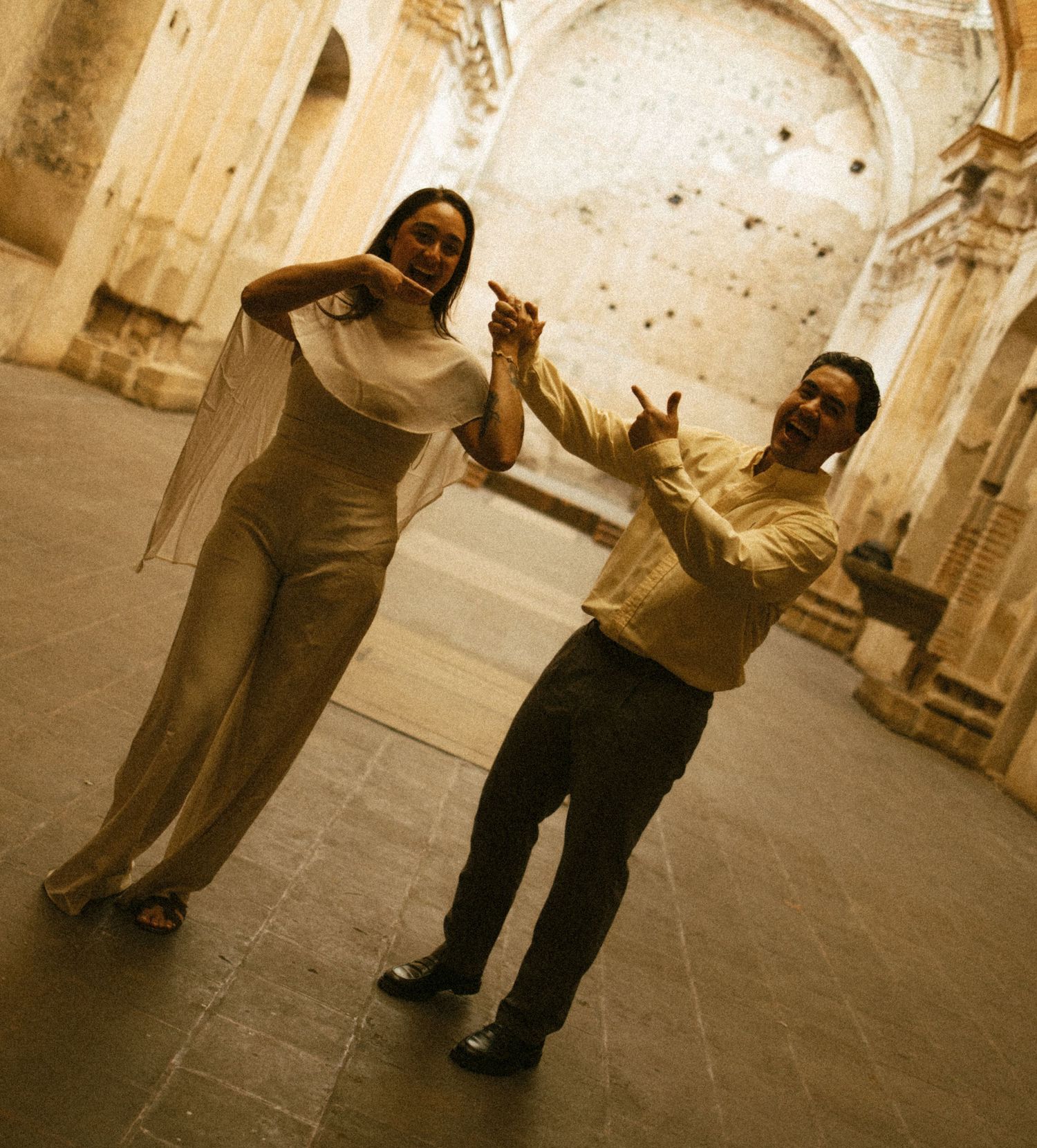 A smiling couple in elegant attire dances hand in hand inside the stone ruins of a historic arched building.