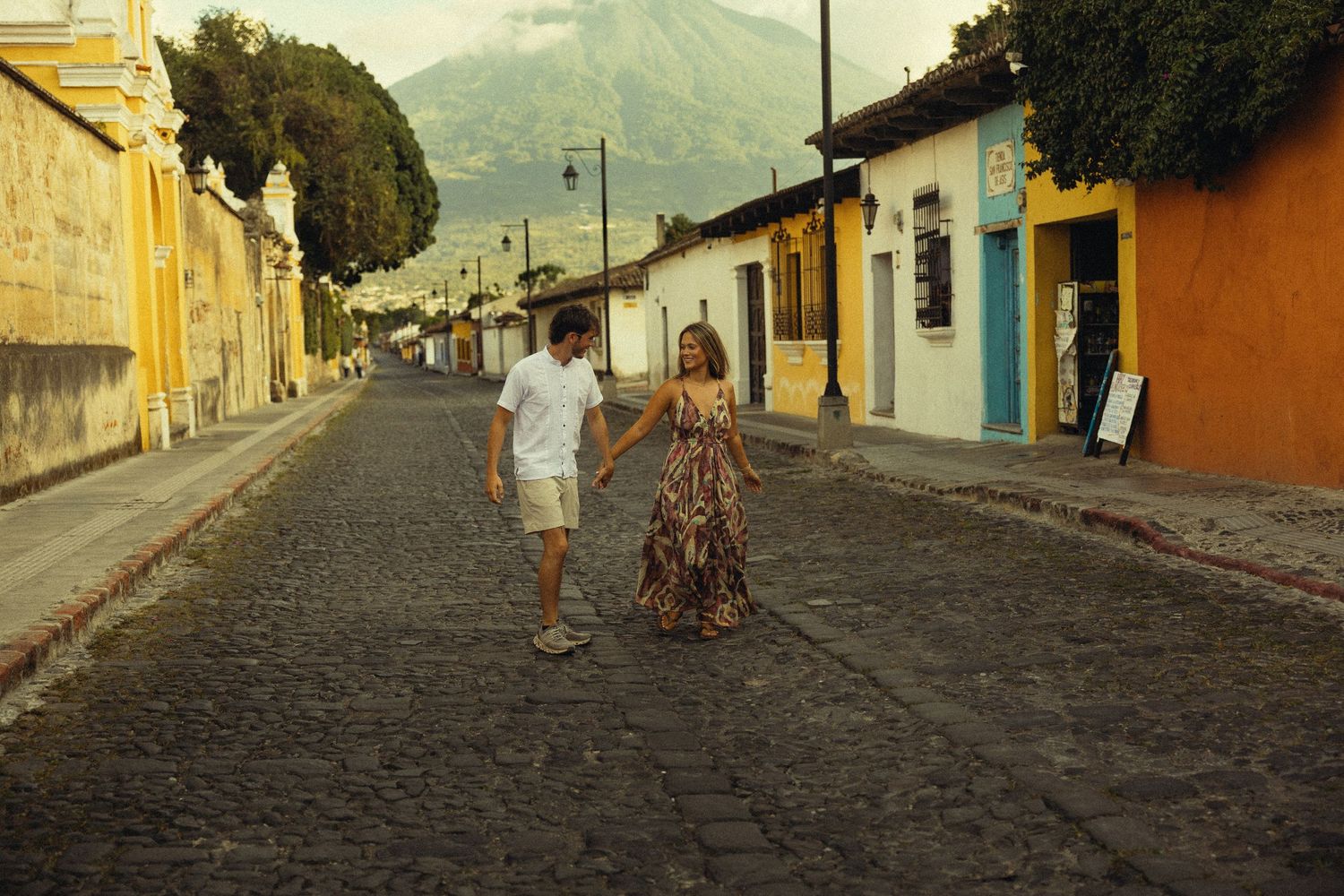 Couple holding hands walking on cobblestone street in Antigua, Guatemala with colorful buildings and volcano backdrop.