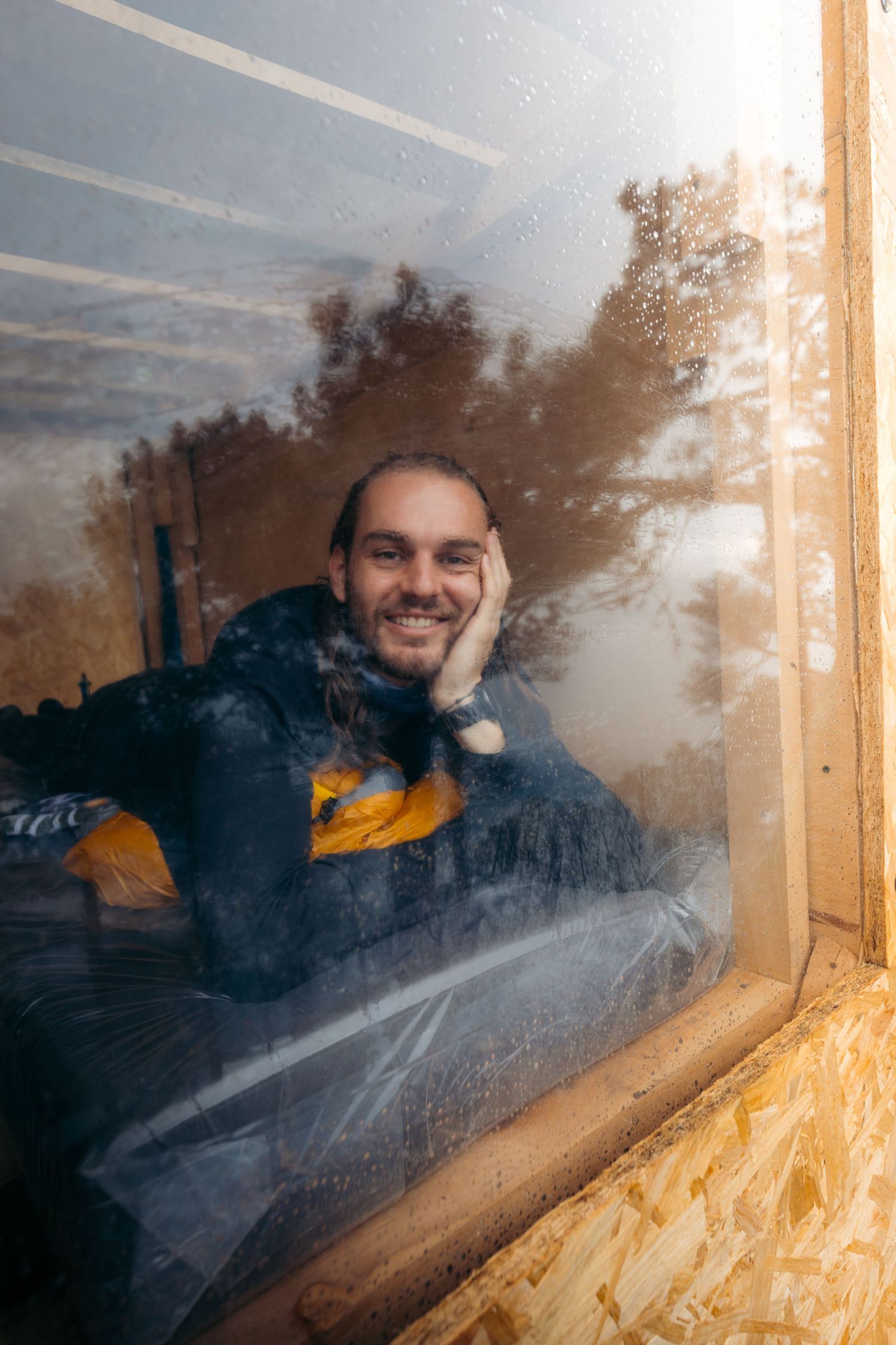 Smiling man with long hair resting his chin on his hand, viewed through a rain-speckled window with tree reflections.