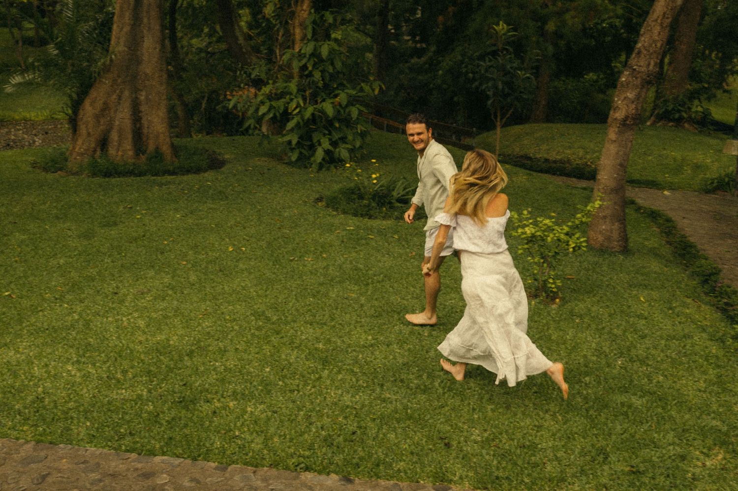 Couple in white outfits running barefoot on lush green grass surrounded by tropical trees.