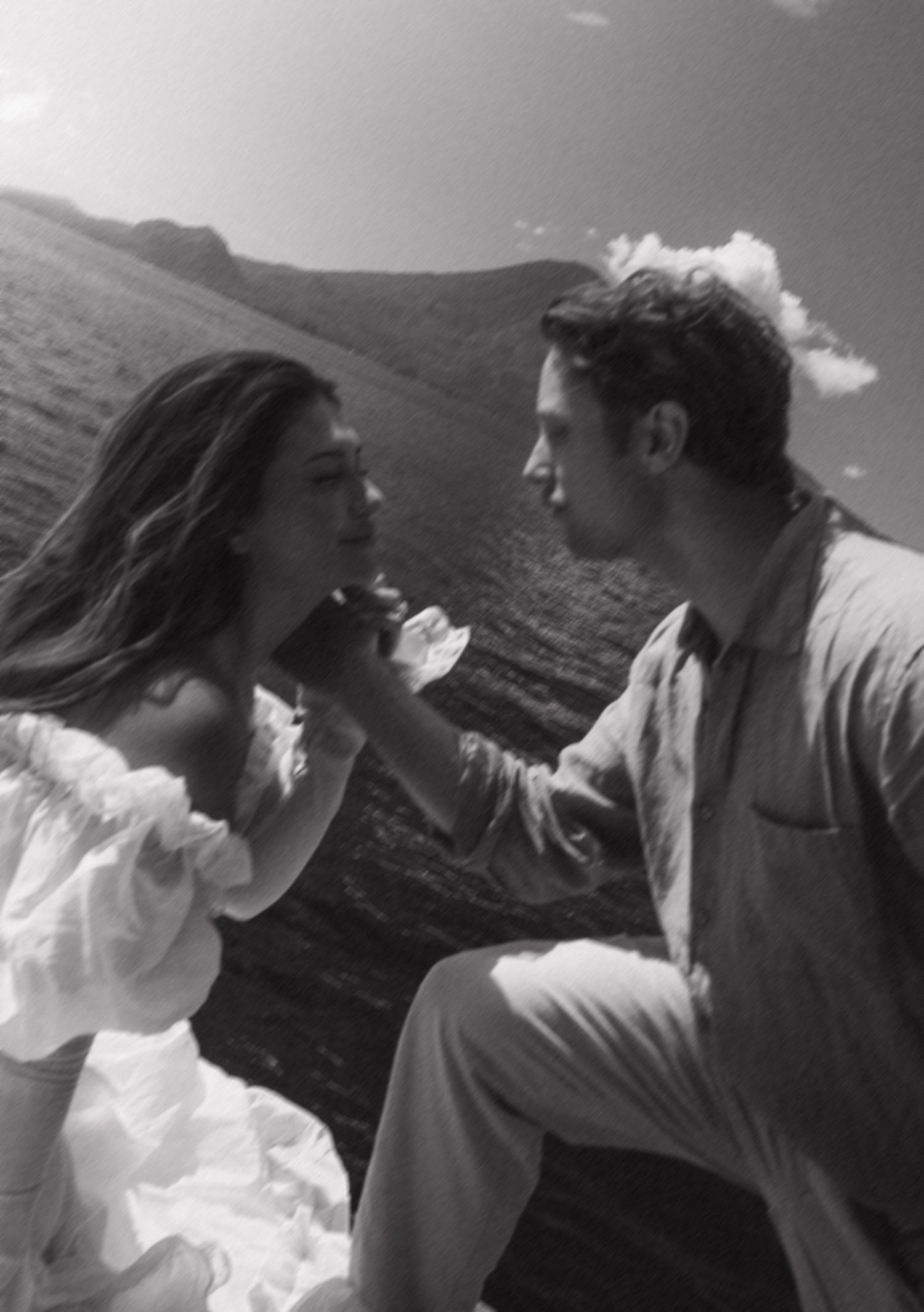 Black and white romantic couple portrait outdoors on a hillside, woman in white off-shoulder dress facing her partner.