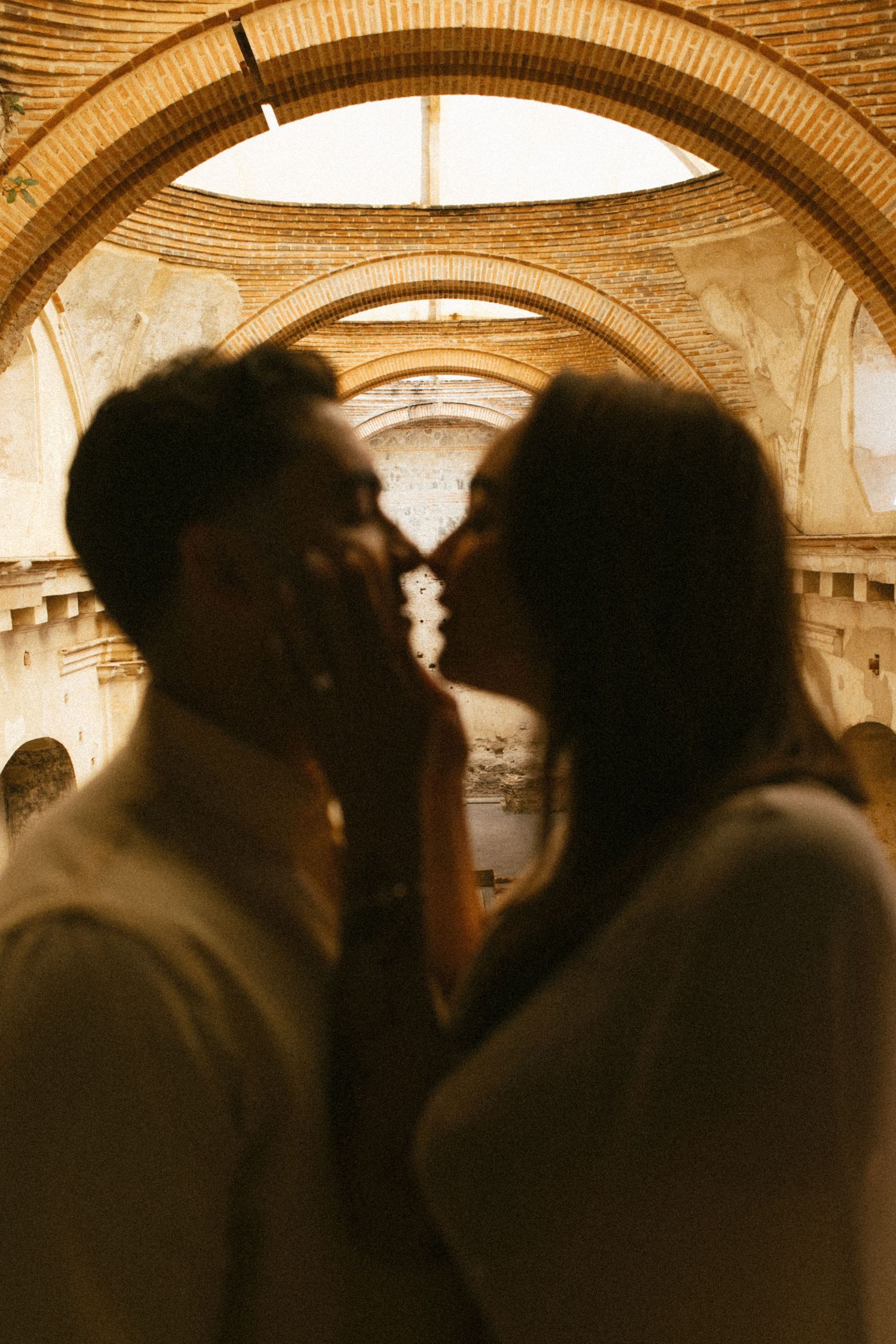 Silhouetted couple sharing a tender kiss inside the warm, golden-lit ruins of an old arched stone building with an open roof.