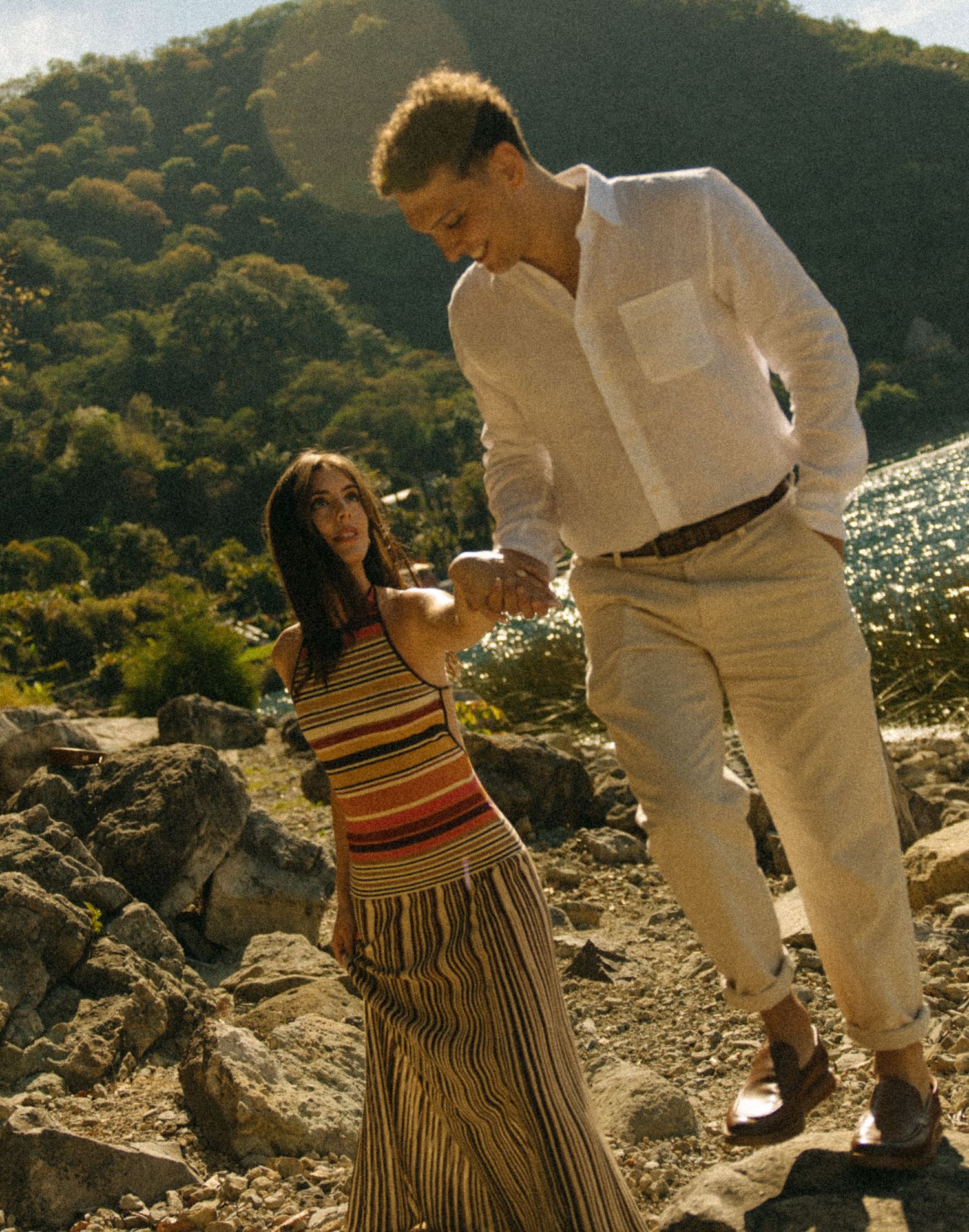 A couple holds hands on rocky terrain, woman in striped maxi dress, man in white linen shirt and cream trousers, green hills behind.