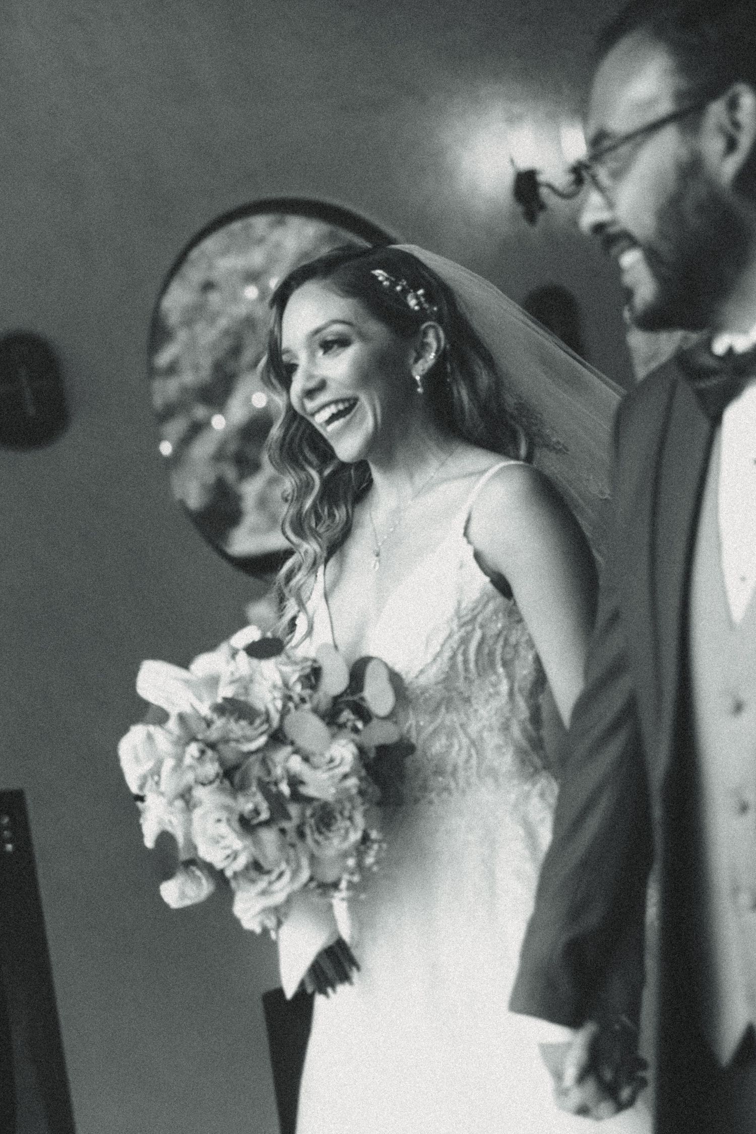 Black and white photo of a smiling bride in a veil holding a bouquet beside a bearded groom in a suit.