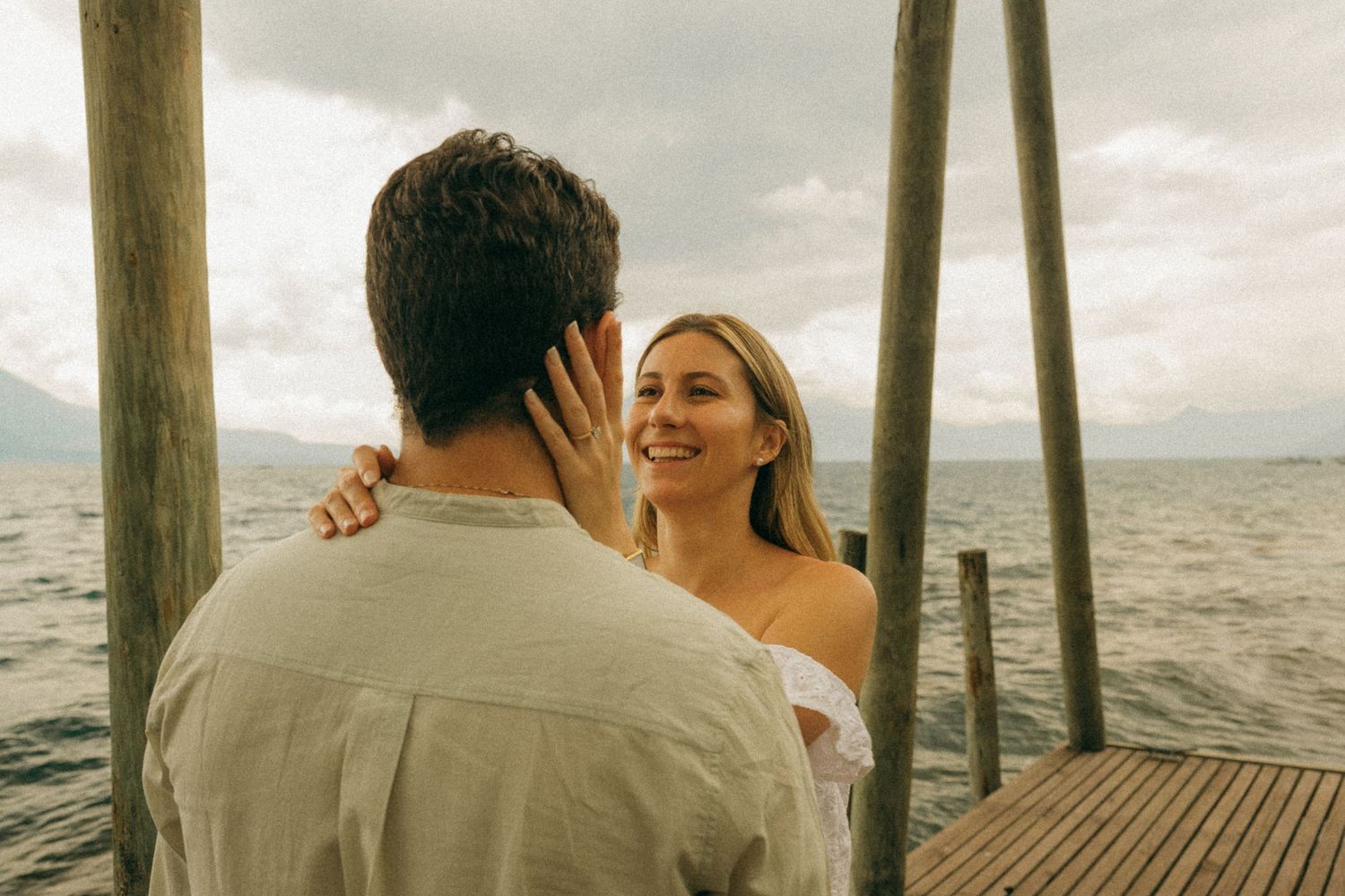 Smiling blonde woman on a wooden dock embraces a man with ocean and cloudy sky in the background.