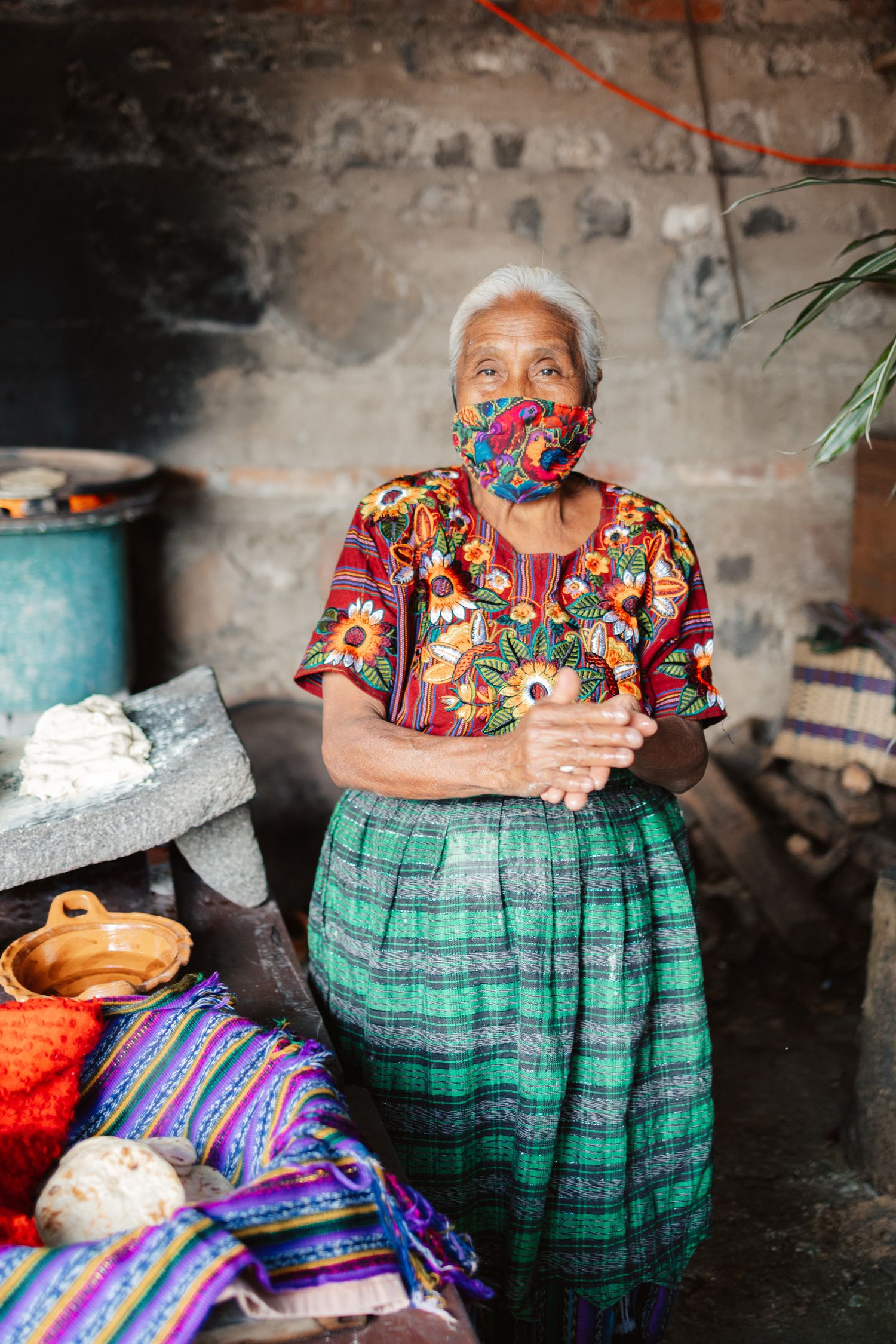 An elderly Guatemalan woman in traditional embroidered huipil and face mask sits in a rustic kitchen.