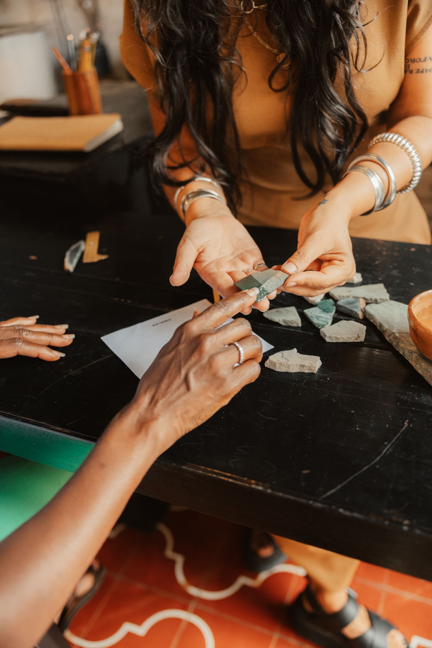 Two women collaborate on a mosaic art project, handling green stone pieces on a dark workshop table.