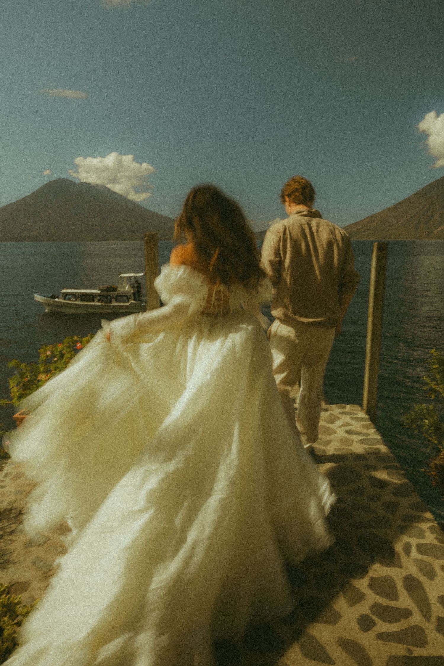 Bride in flowing white gown and groom walk hand-in-hand on a stone pier overlooking a volcanic lake.