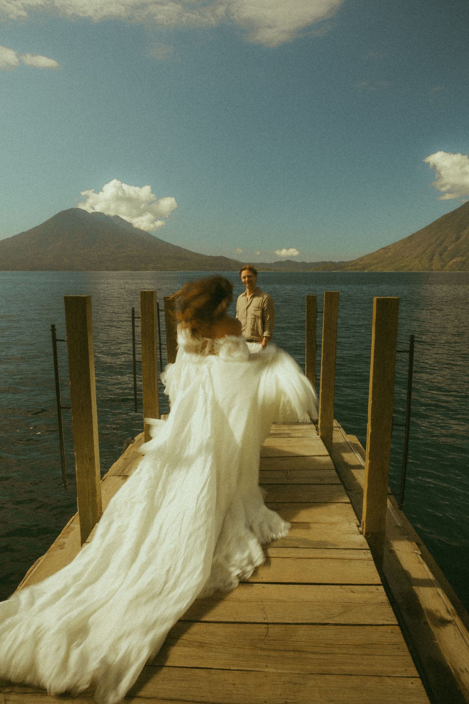 Bride in flowing white gown walks toward groom on wooden dock over lake with volcanic mountains backdrop.