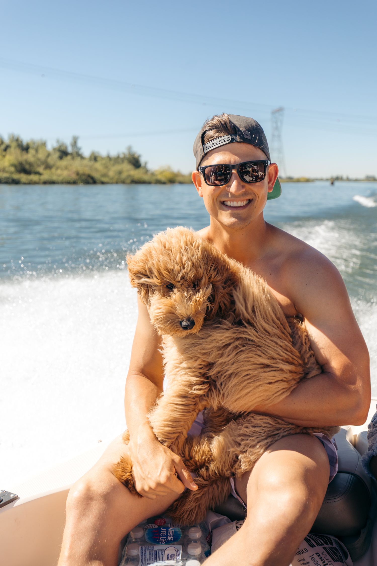 Smiling shirtless man holding a fluffy golden doodle puppy on a speedboat with green trees and blue sky.