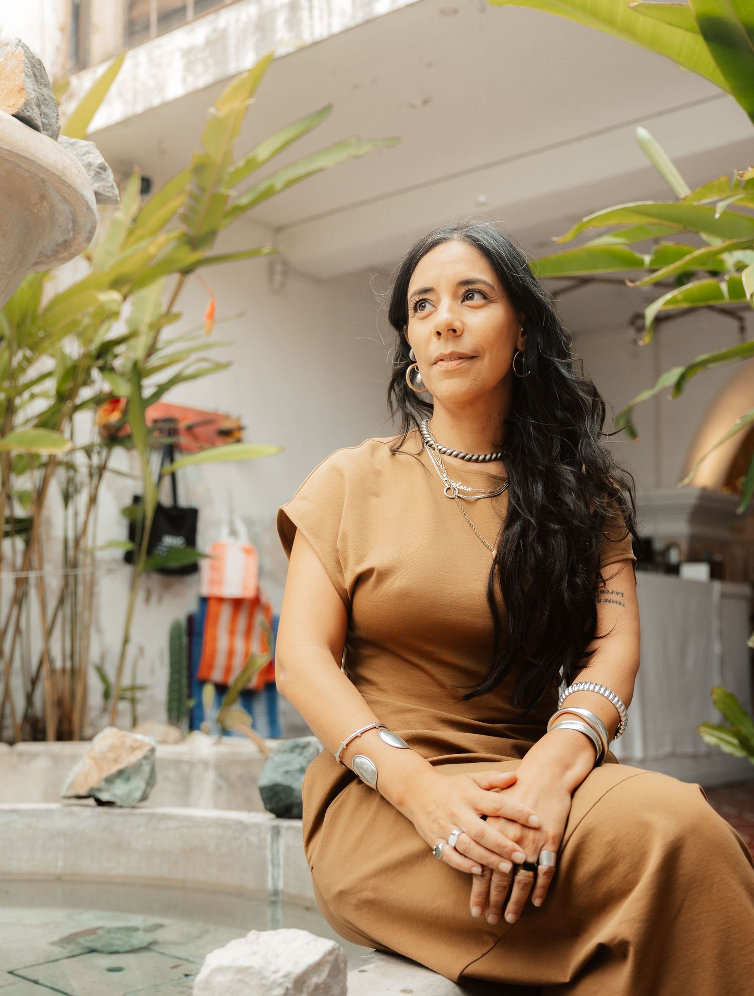 Woman in tan dress with silver jewelry sits outdoors near fountain surrounded by tropical plants.