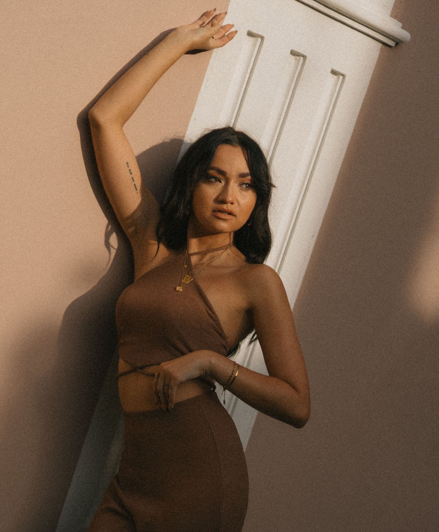 Woman in brown cutout dress posing against white door in warm golden light, wearing gold jewelry.