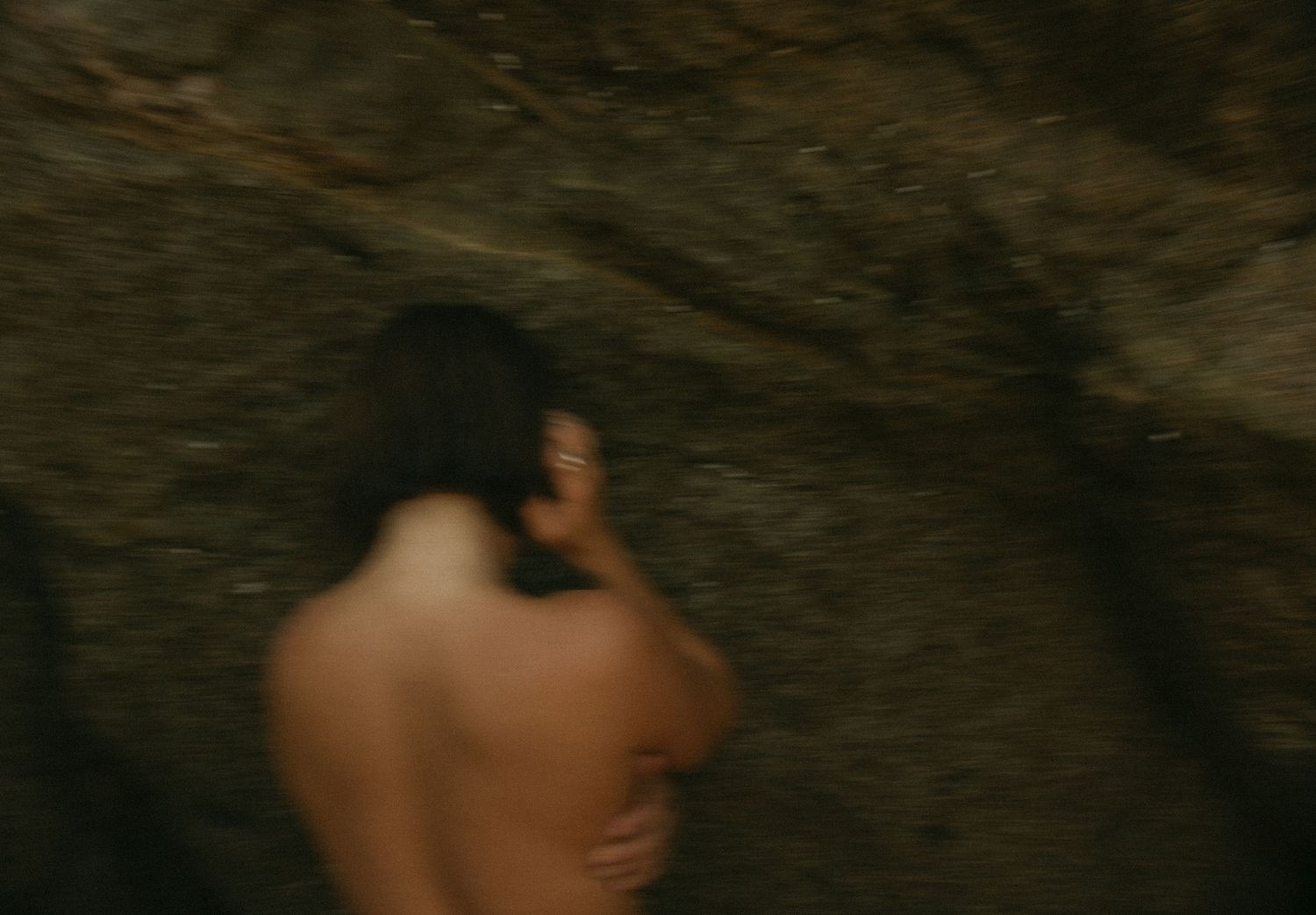 A blurred figure with dark hair stands against a rocky surface in dim, moody lighting.