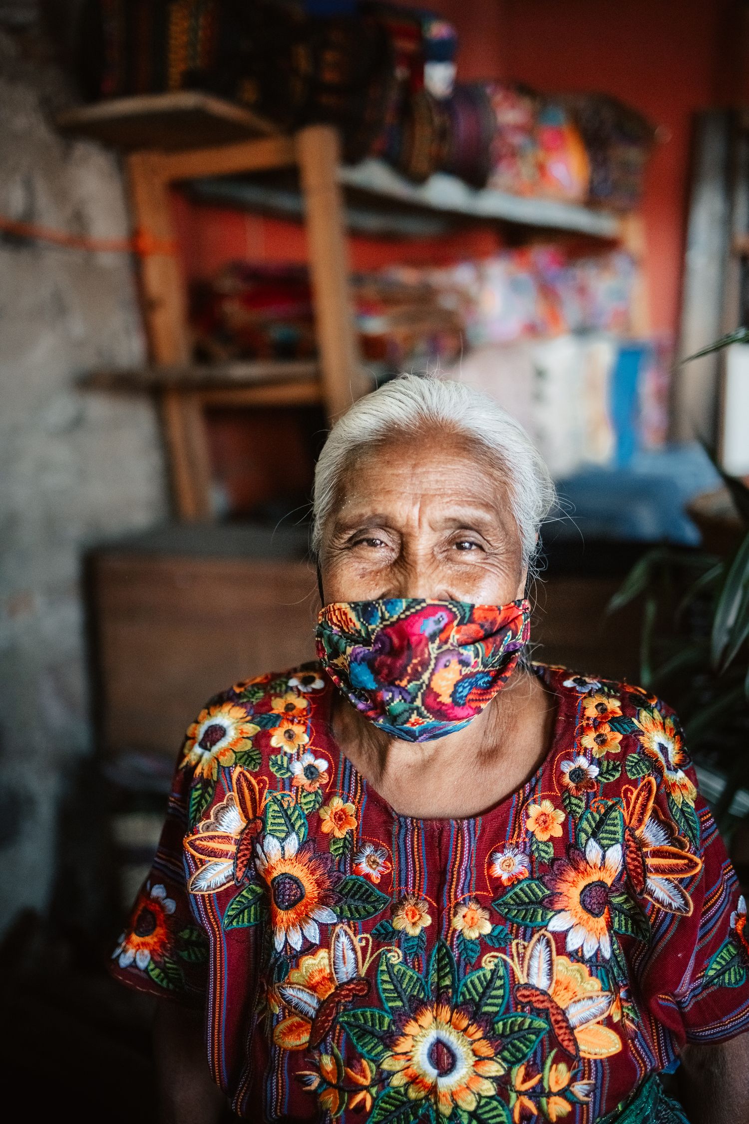 Elderly indigenous woman in vibrant floral dress and matching fabric face mask stands in a colorful shop.