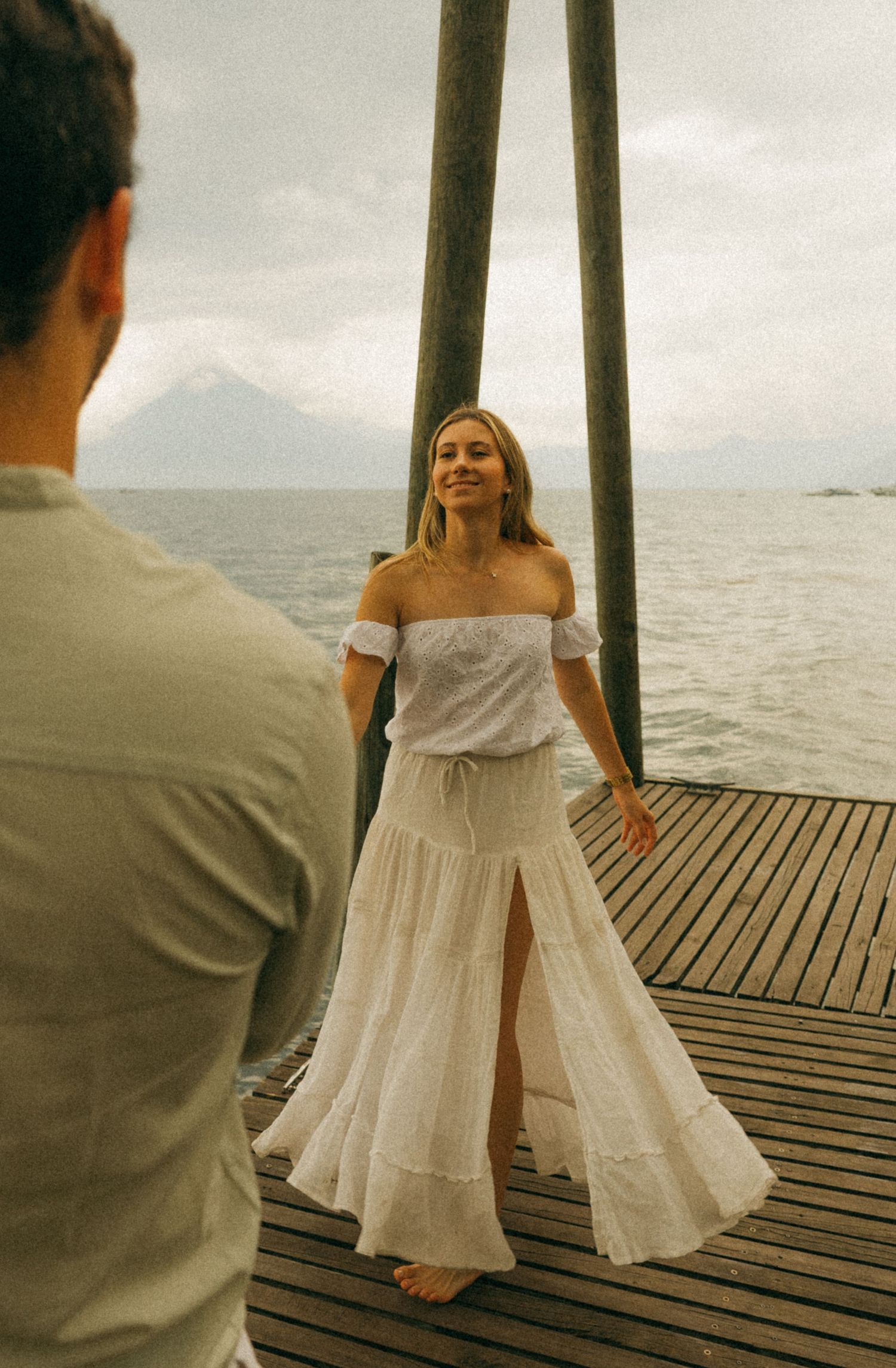 Woman in white off-shoulder top and maxi skirt standing on wooden dock overlooking lake with volcano.
