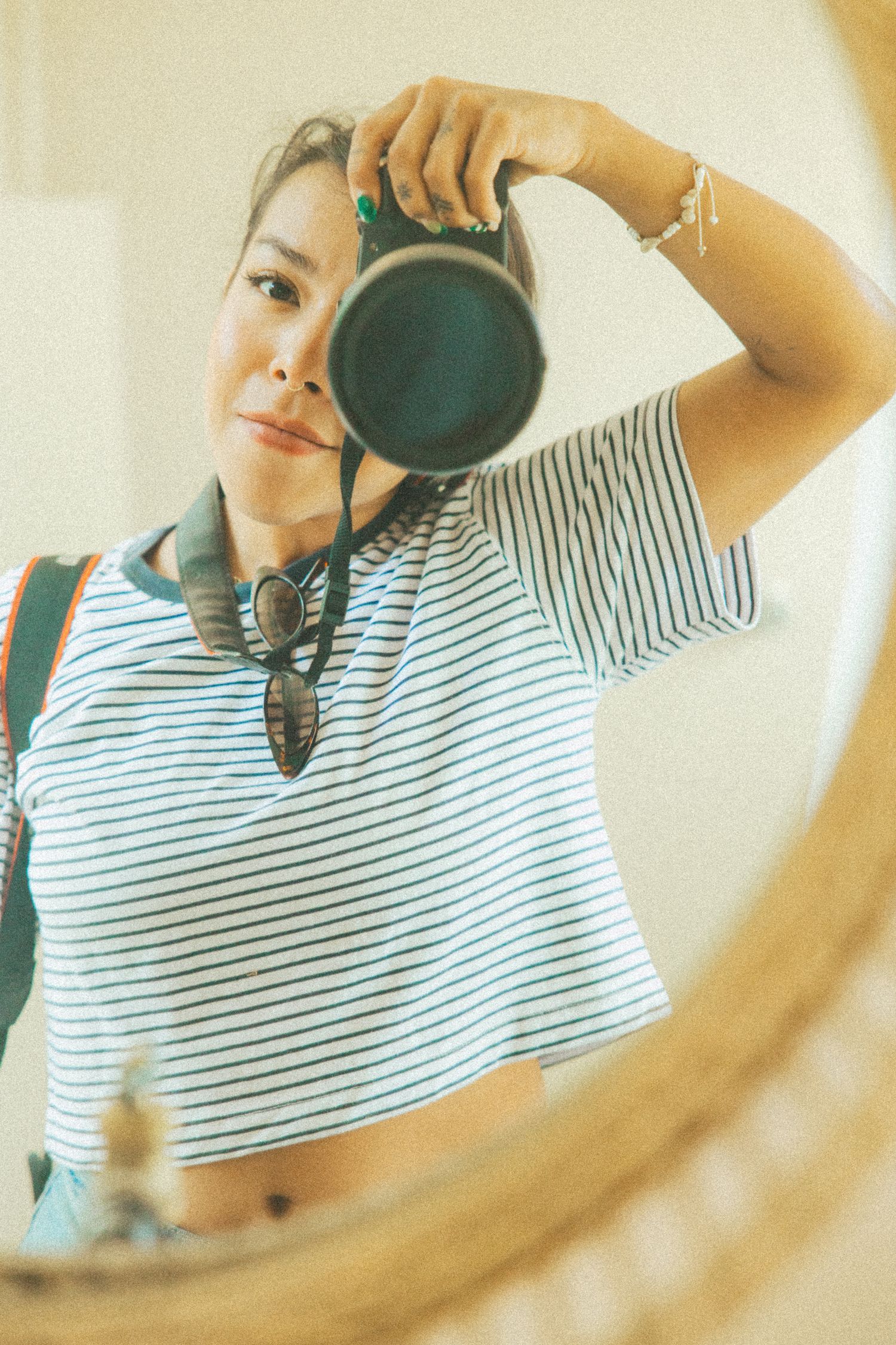 Woman in striped shirt takes mirror selfie with DSLR camera, wearing sunglasses around neck and bracelet.