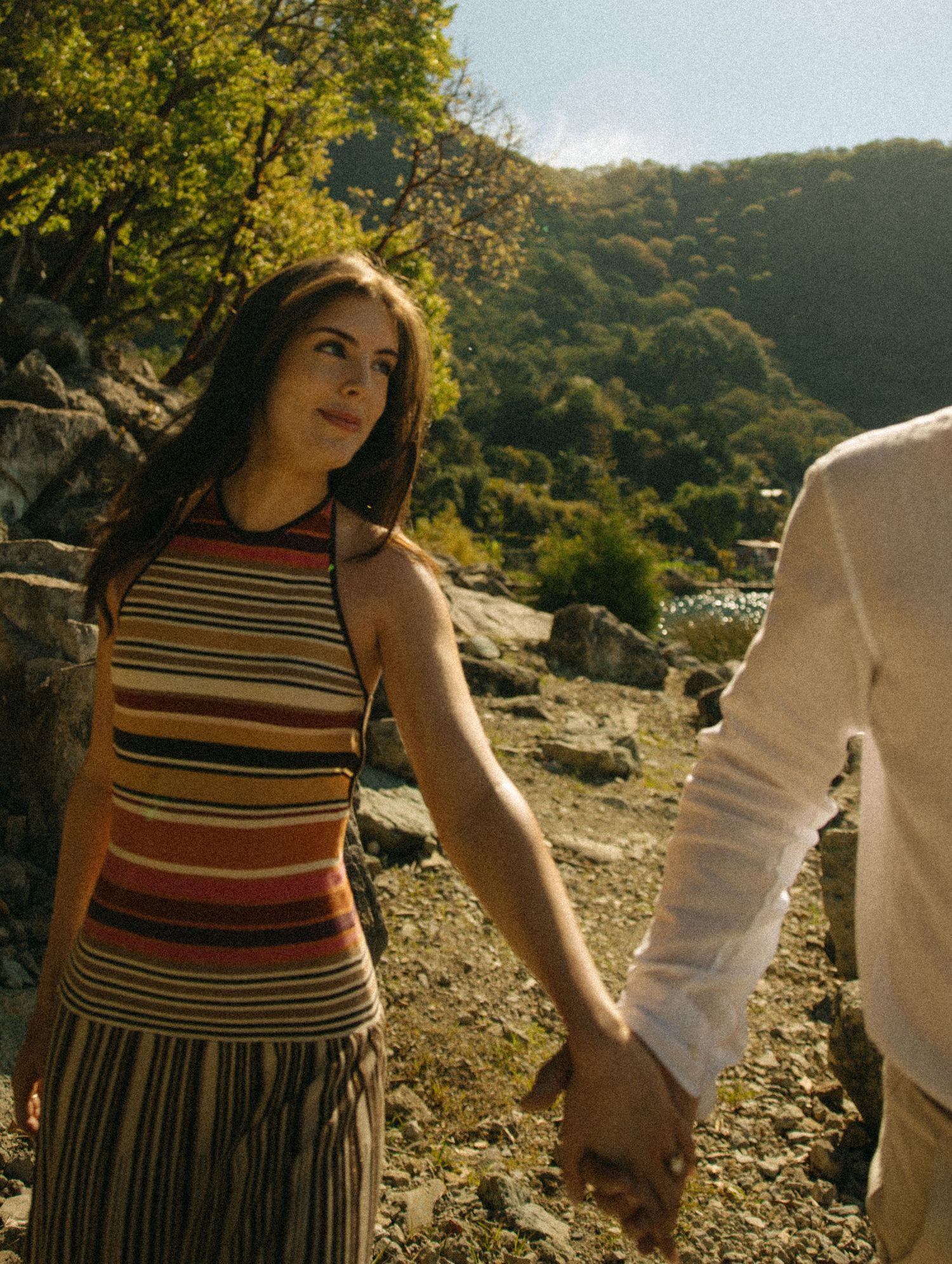 A woman in a striped halter top holds hands with a partner outdoors near rocky terrain and lush green hills.