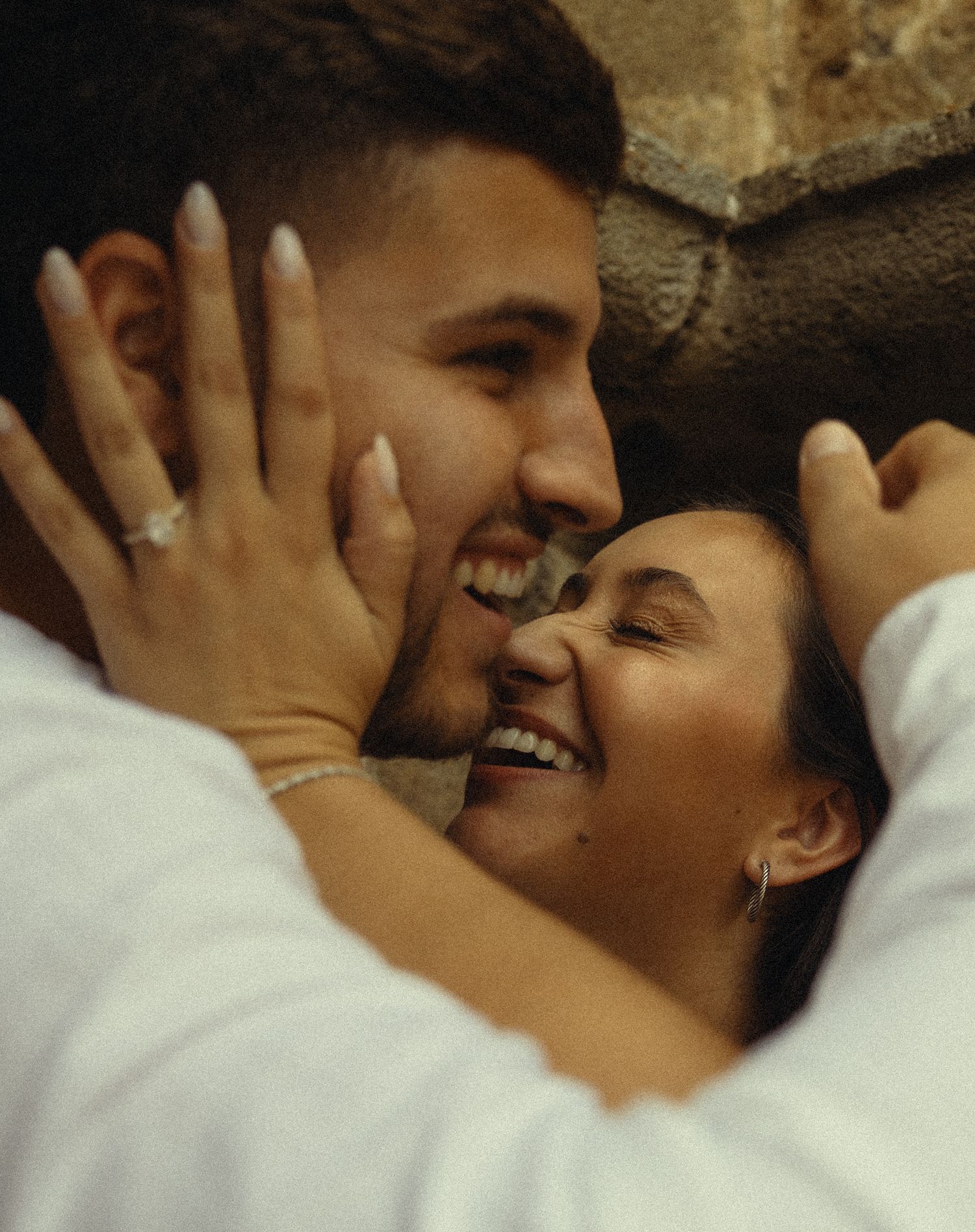Happy couple embracing and smiling, woman wearing engagement ring and bracelet, romantic moment near stone wall.