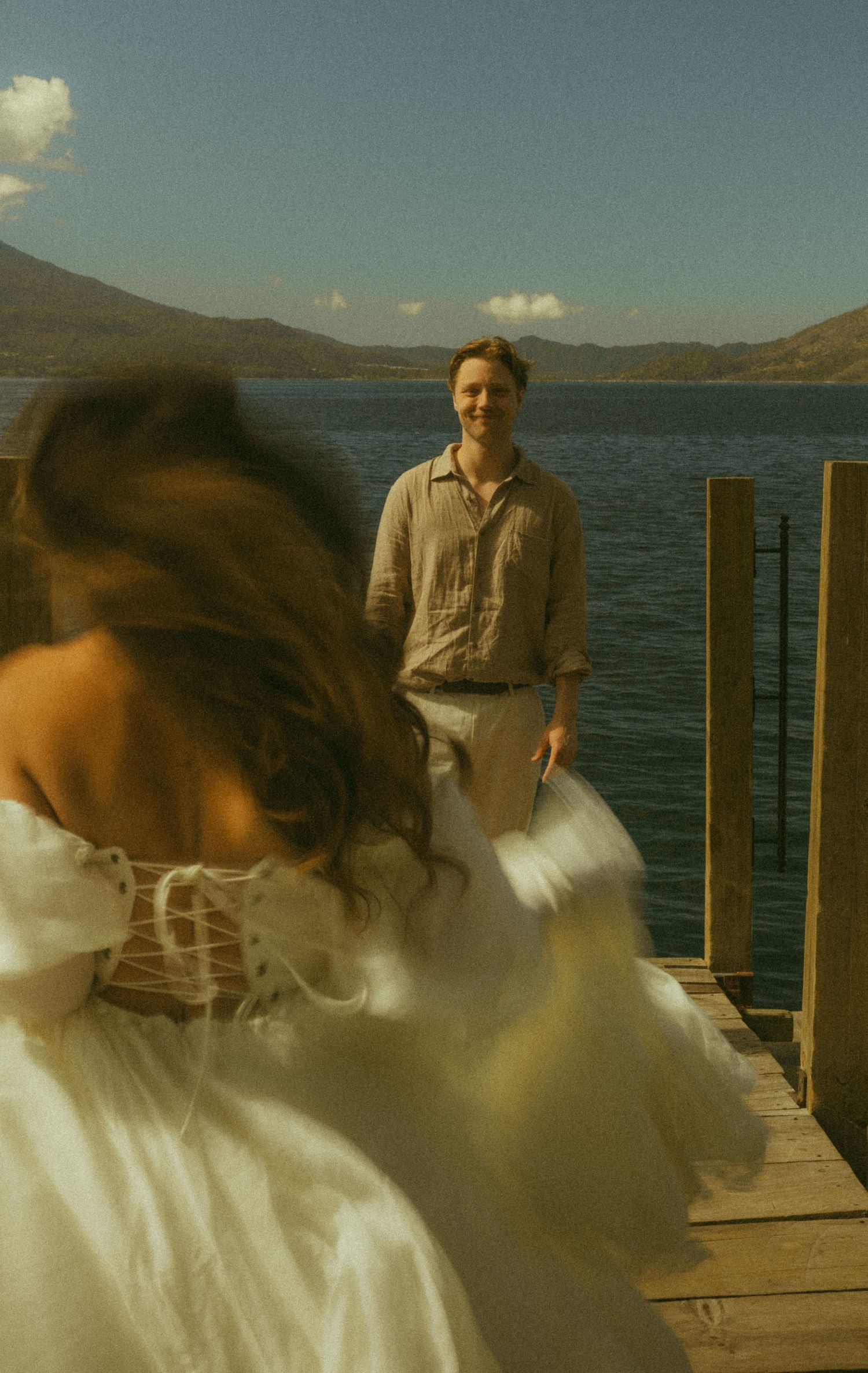 Bride in white corset gown walking toward smiling groom on wooden dock overlooking mountain lake.