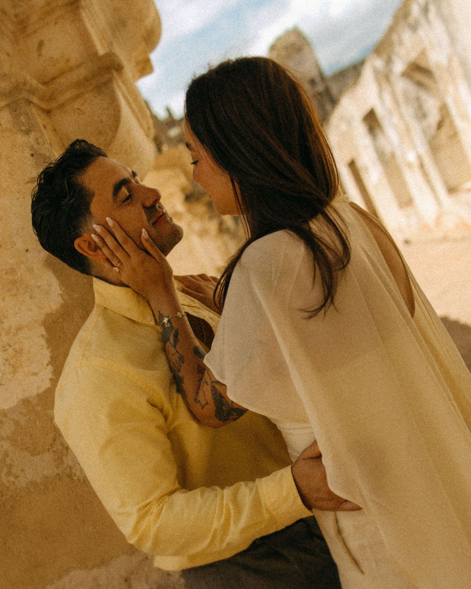 Romantic couple sharing tender moment leaning against ancient stone column, man with tattoos in yellow shirt.