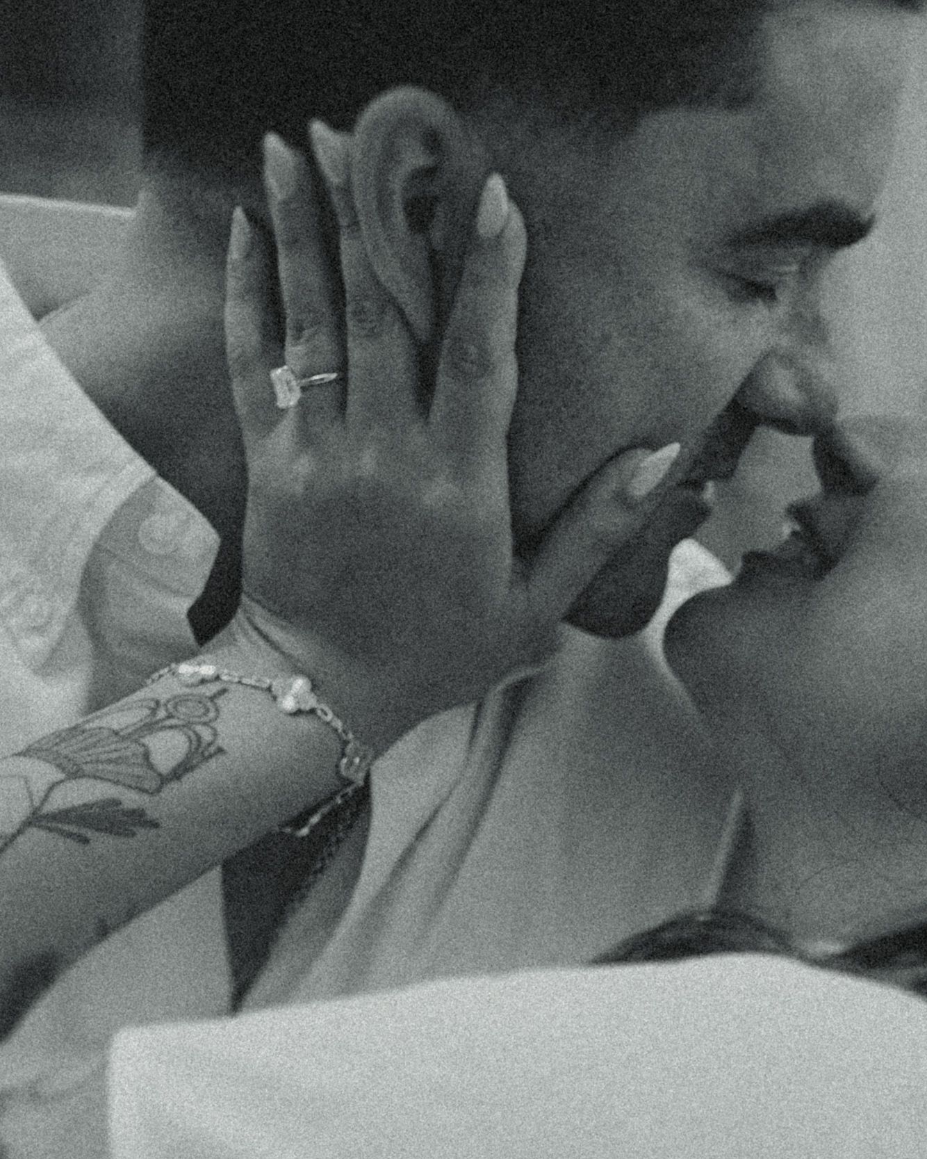 Black and white close-up of a couple about to kiss, her hand with a diamond ring and floral tattoo cradling his face.