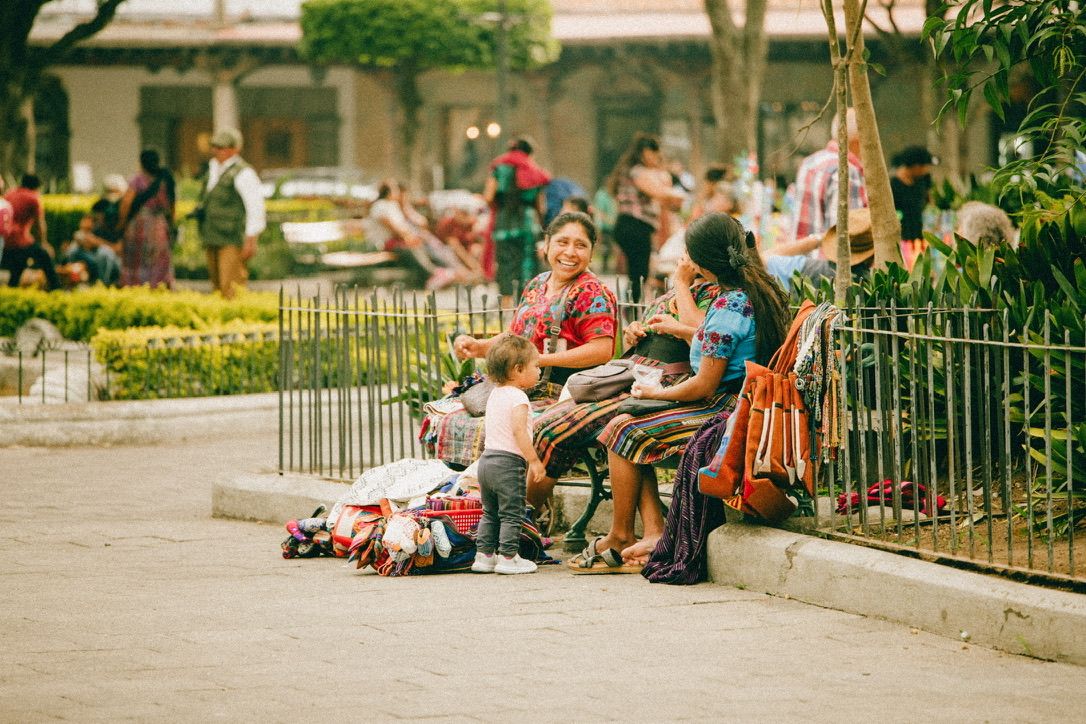 Women and children sitting on a bench in a bustling outdoor market plaza in Guatemala.