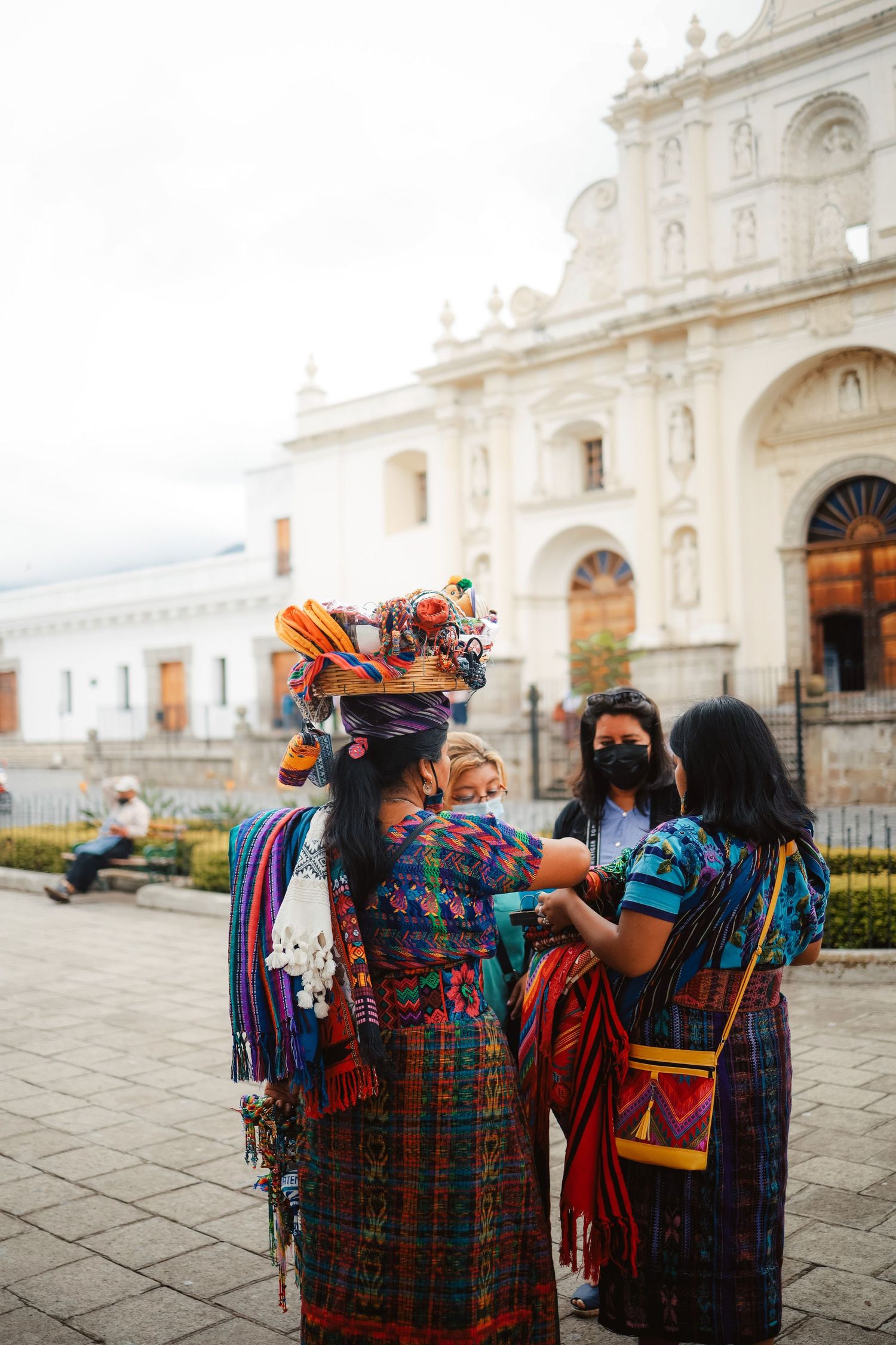 Indigenous Guatemalan women in colorful traditional traje clothing near a white colonial church in Antigua, Guatemala.