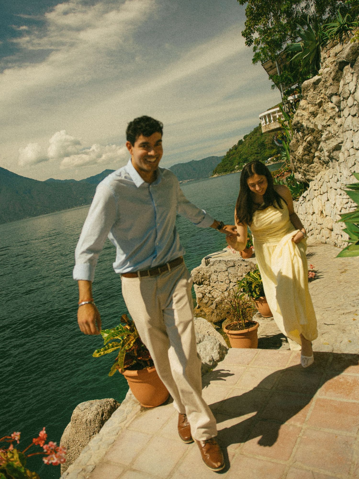 Smiling couple walks hand in hand along a terracotta lakeside terrace with potted plants, mountains and cloudy sky in the background.