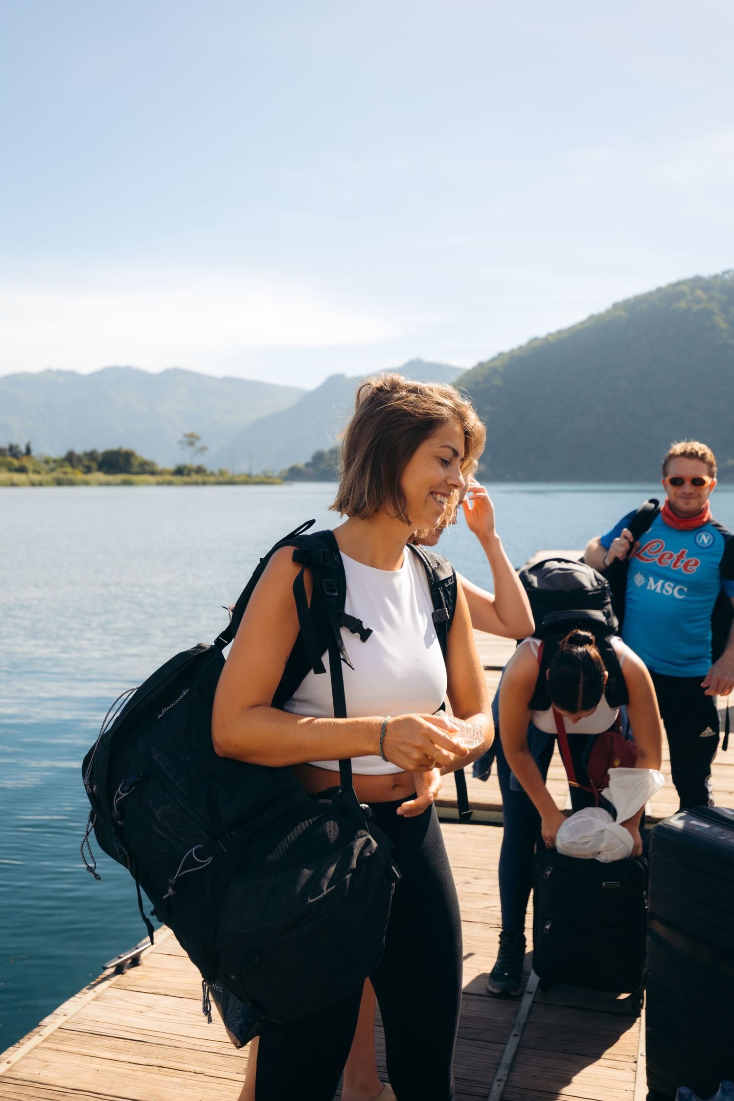 Young woman with backpack smiling on a wooden dock by a lake, with mountains and blue sky in the background.