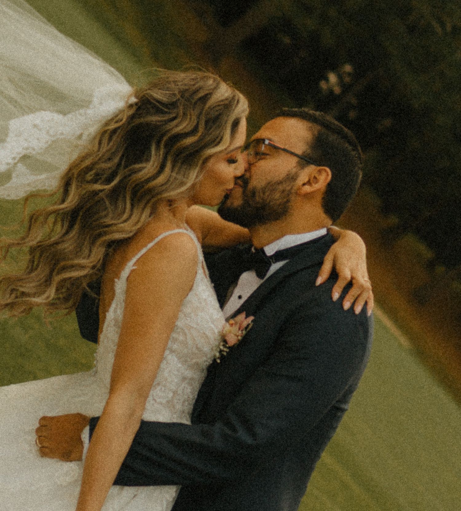 A bride and groom share a romantic kiss outdoors, the bride in a white lace gown with flowing hair.