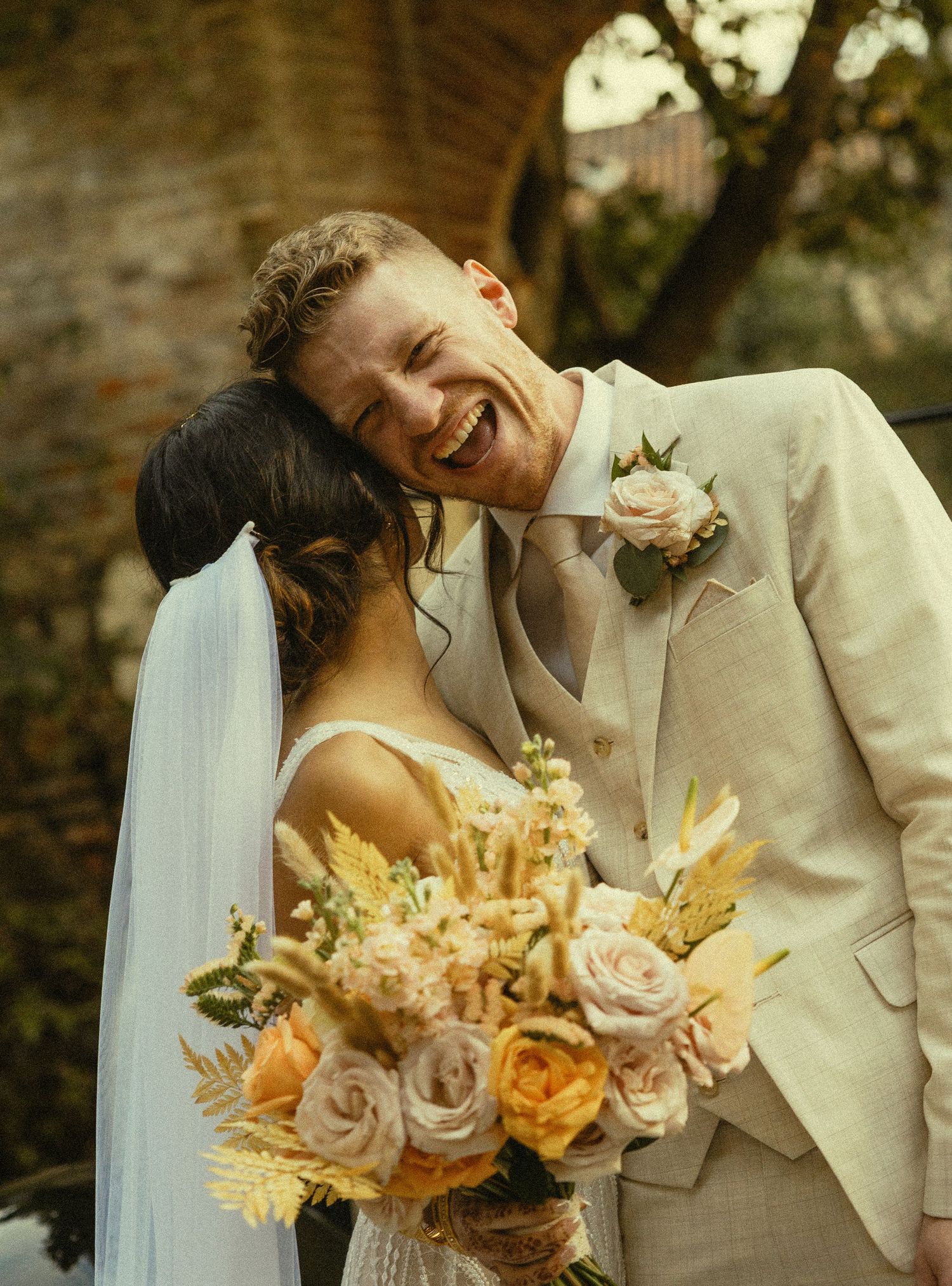 Joyful groom in beige suit laughing as bride in white veil holds colorful bouquet of roses at wedding.