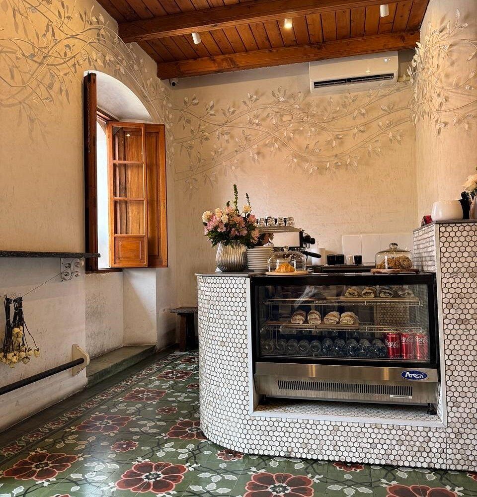 Charming cafe interior with penny tile counter, vintage floor tiles, ornate walls, display case with pastries, and wooden ceiling.