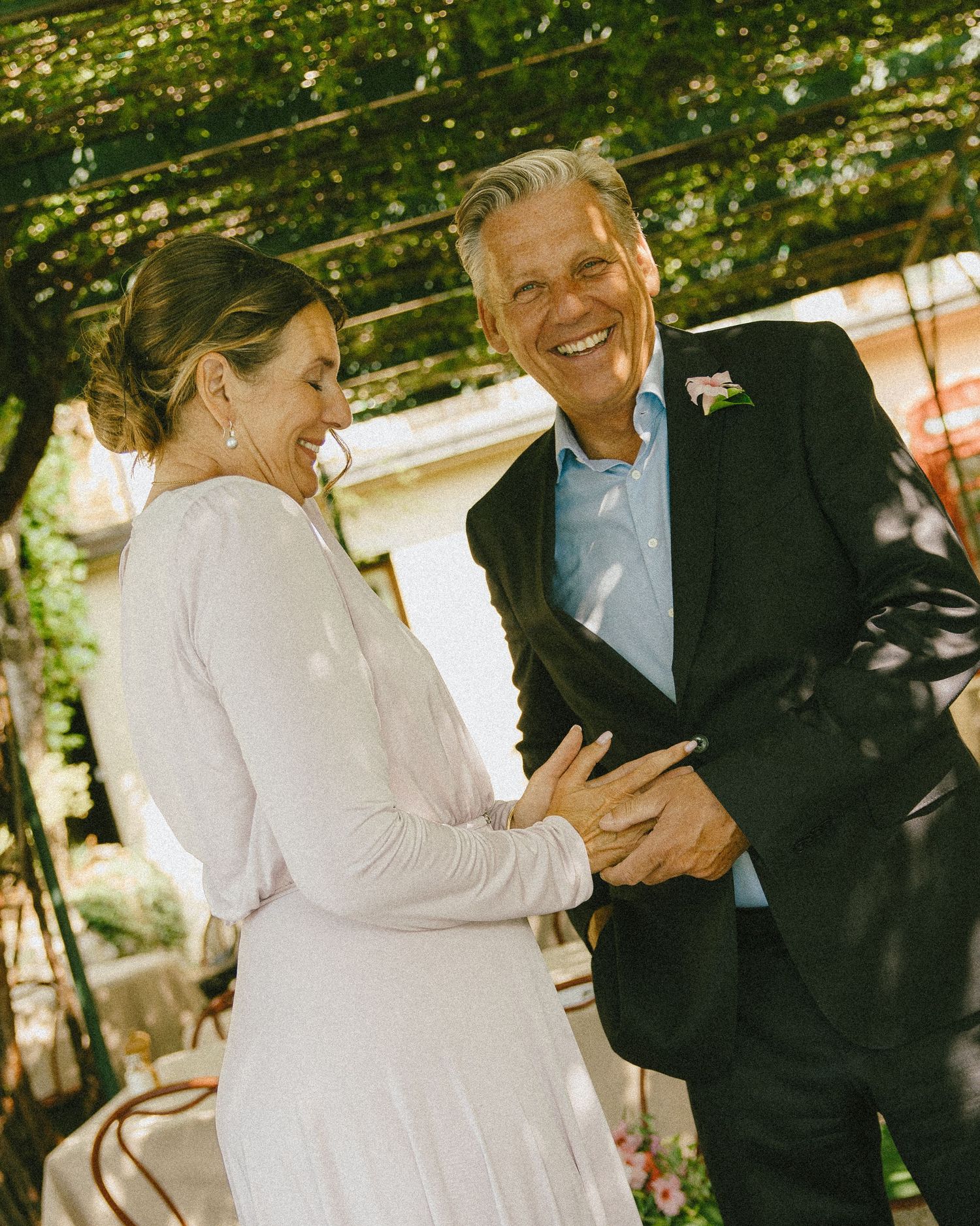 Couple laughing joyfully during outdoor wedding ceremony under green vine-covered pergola, bride in white dress, groom in dark suit.