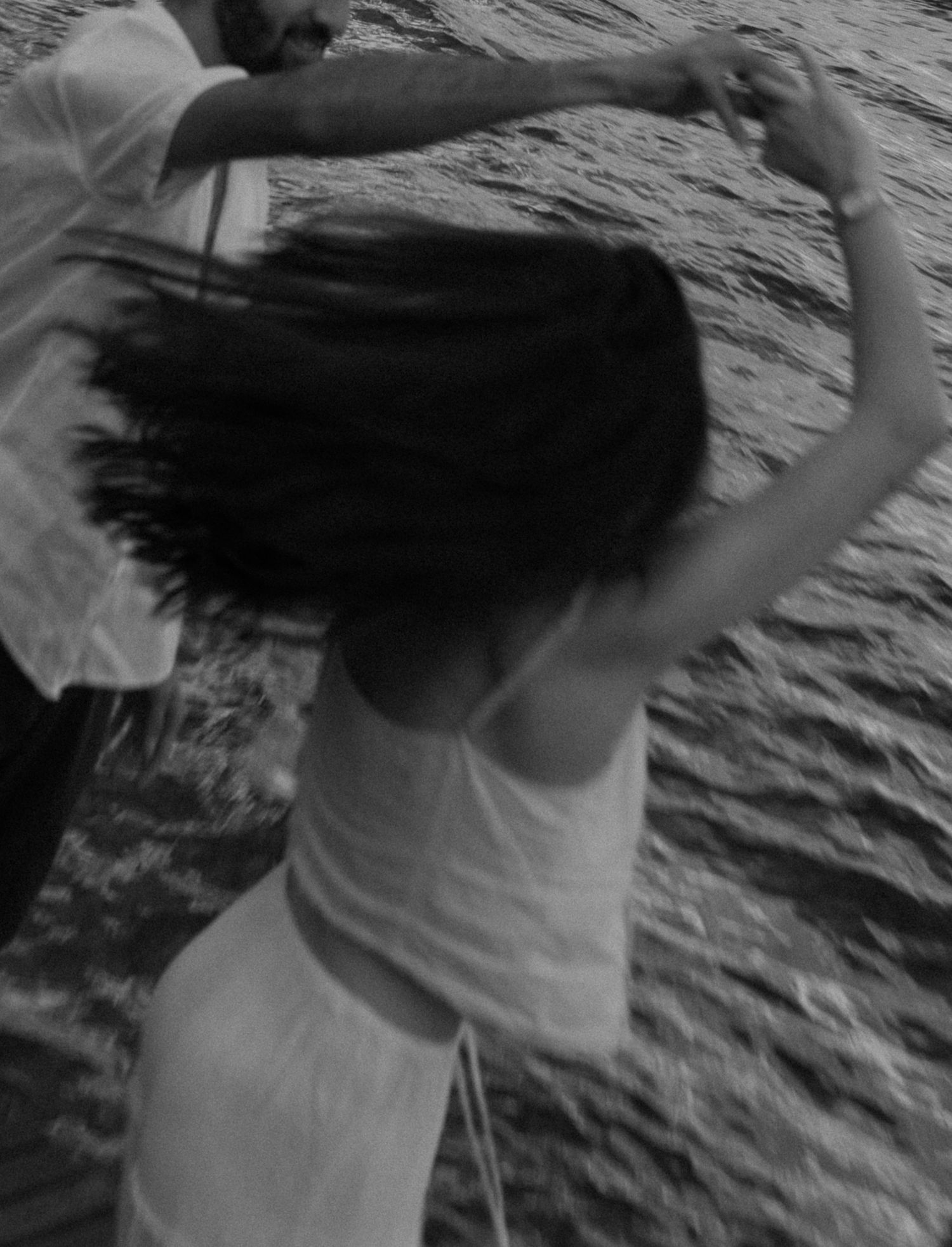 Black and white photo of a couple dancing near water, woman in white dress with flowing dark hair.