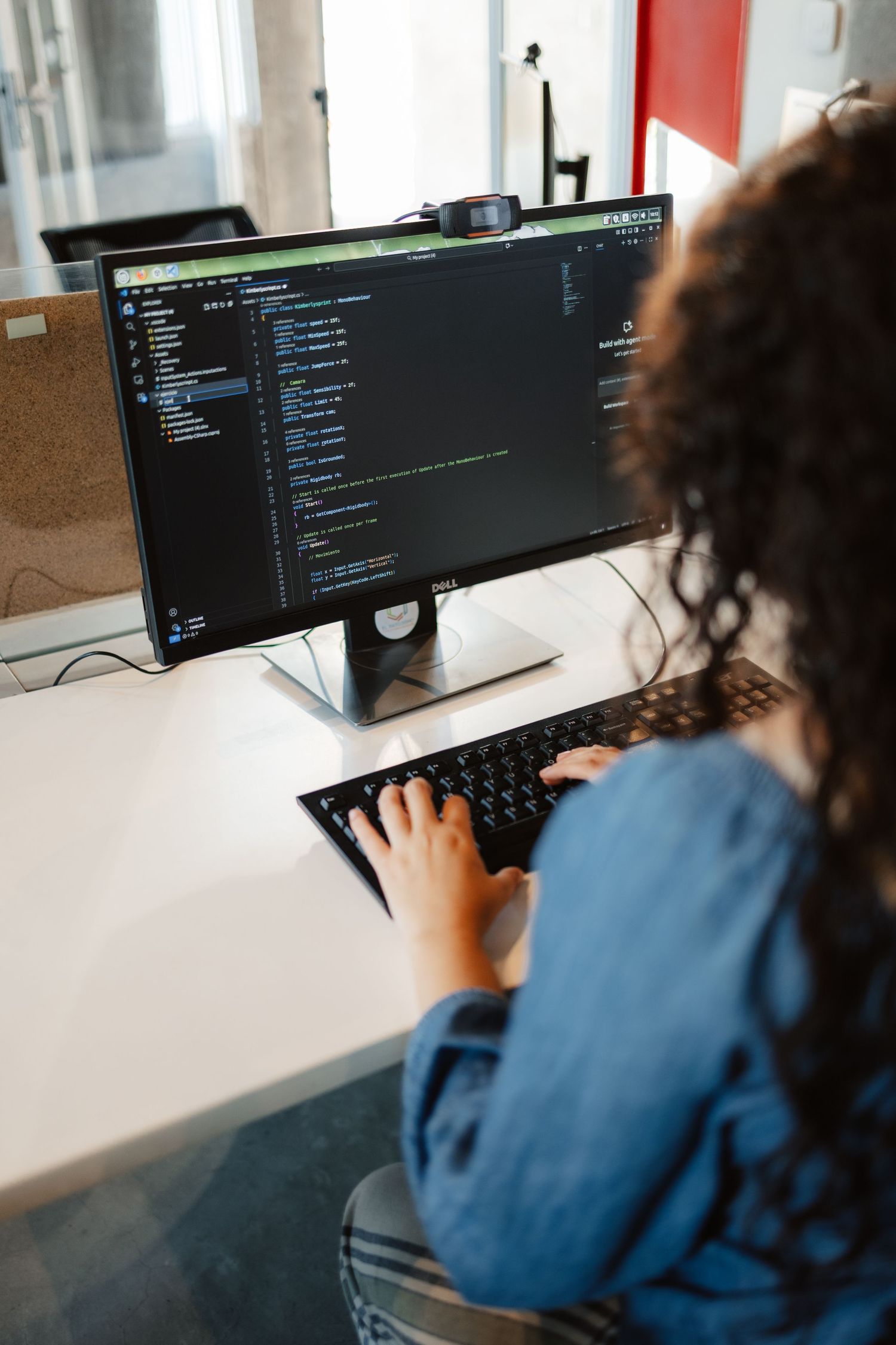 Woman with curly hair coding on a dark-themed IDE on a Dell monitor in a modern office.