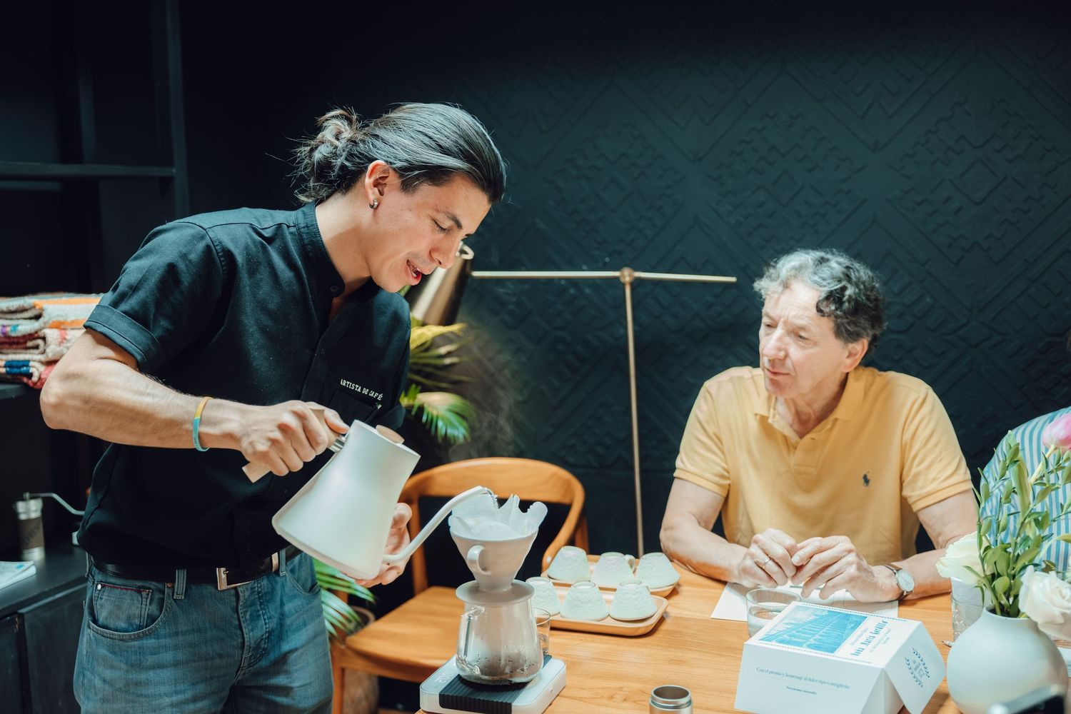 Barista pouring hot water into a pour-over coffee dripper for a seated guest at a wooden cafe table.