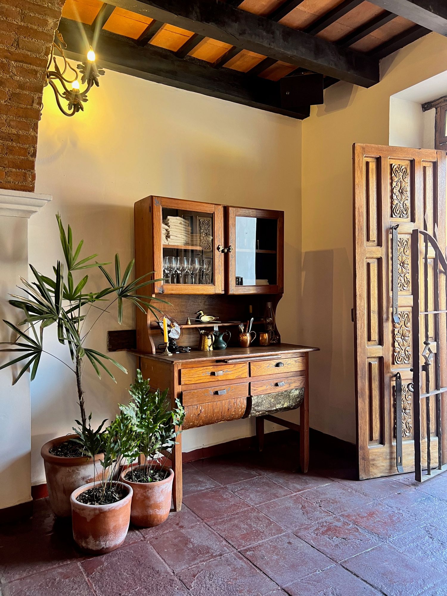 Rustic Tuscan interior with antique wooden hutch, terracotta tile floor, potted plants, and ornate carved wooden door.