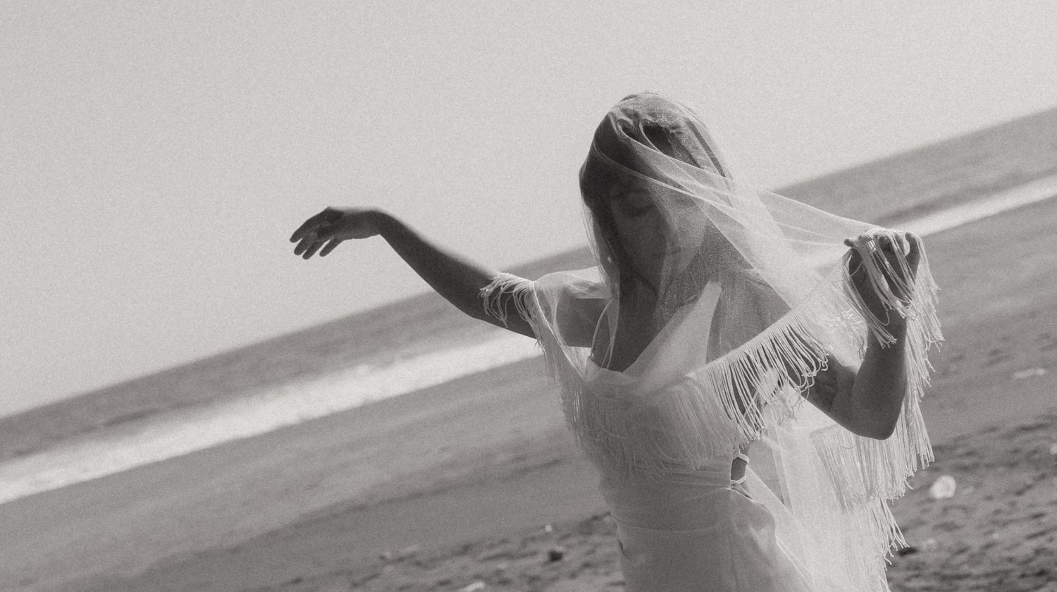 Black and white photo of a bride in a white dress and veil posing gracefully on a beach.