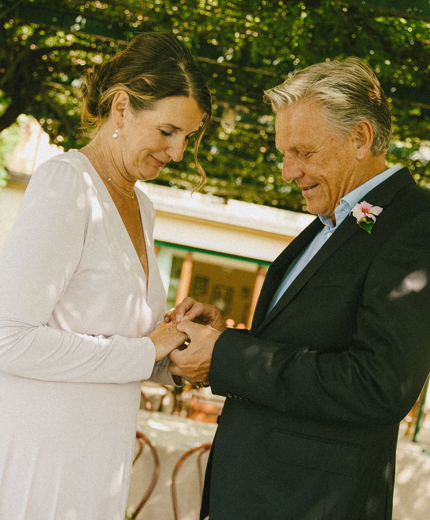 Smiling bride in a white dress and groom in a dark suit with a pink boutonniere exchange rings during an outdoor garden wedding ceremony.