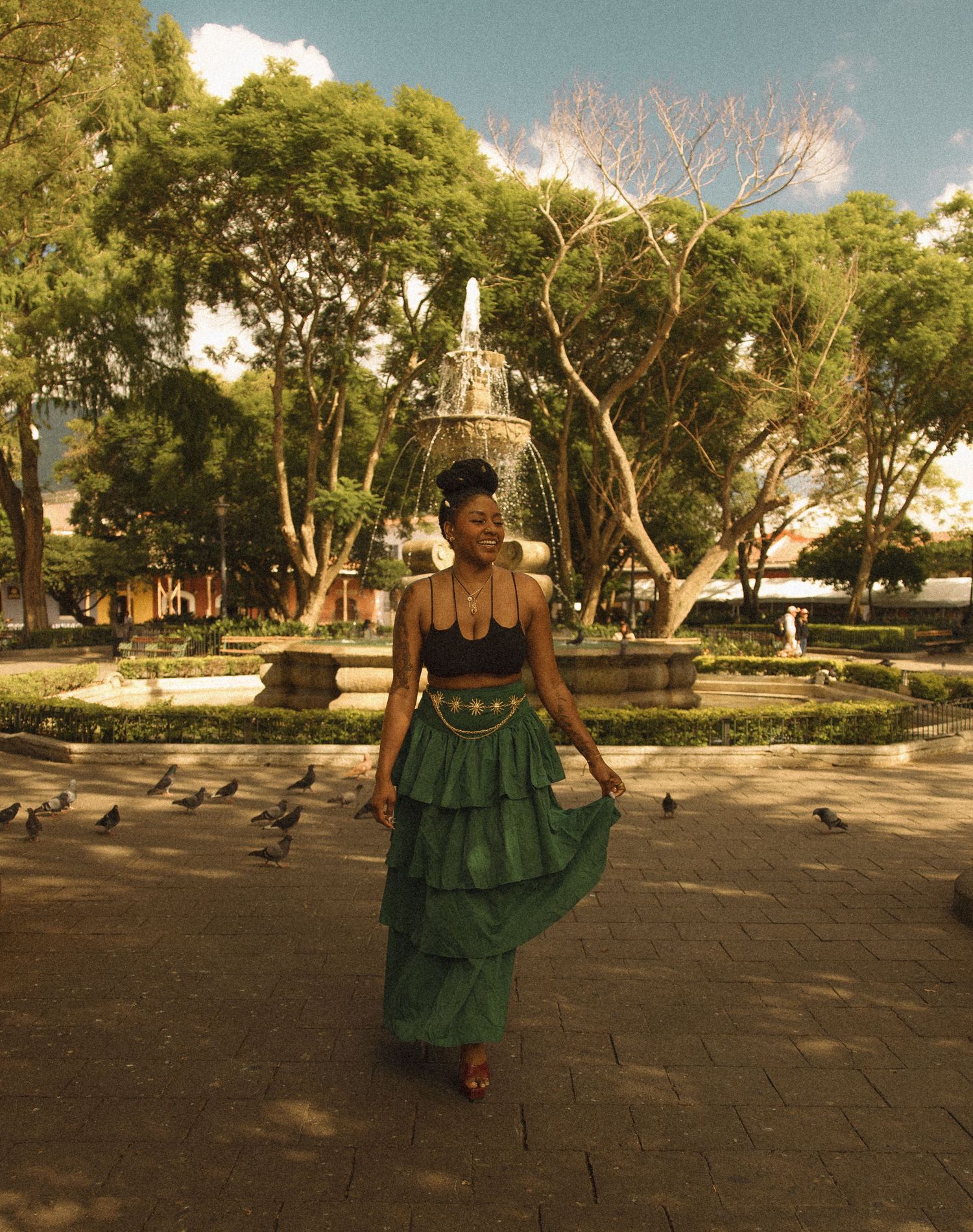 A woman in a green ruffled skirt and black top walks through a plaza with a fountain and trees.